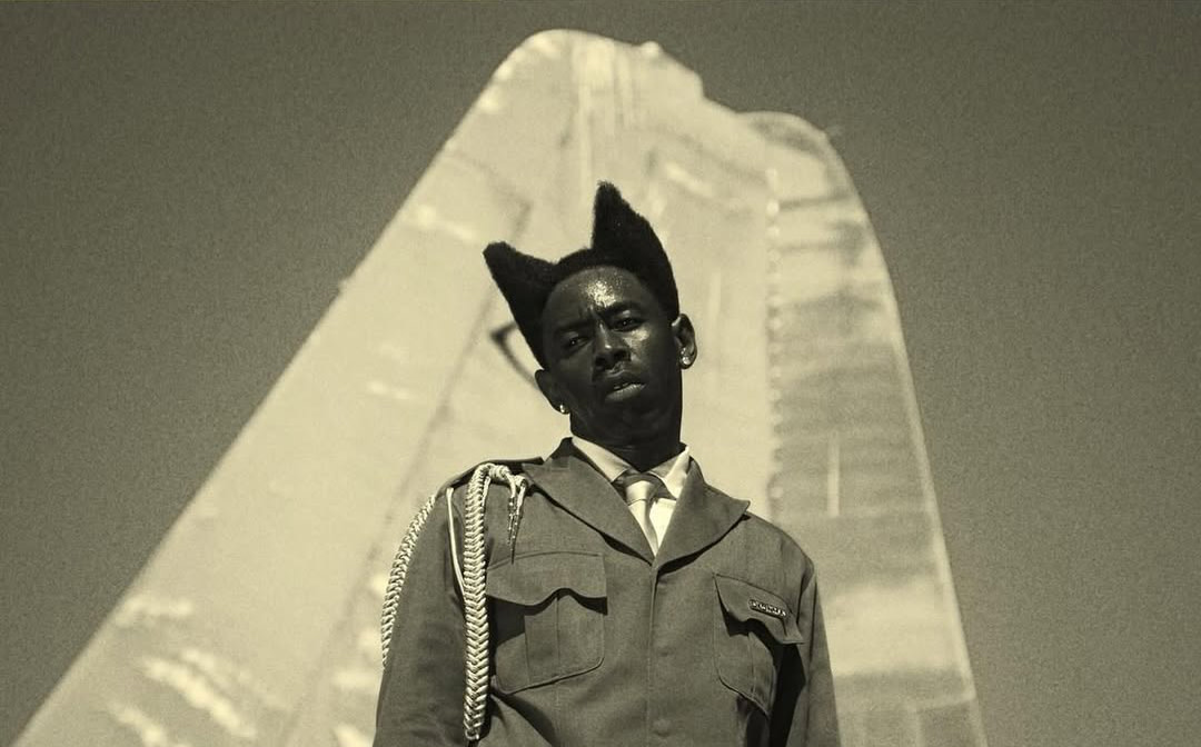 Tyler, the Creator in a suit with unique hair stands in front of a large, abstract structure in a sepia-toned image.