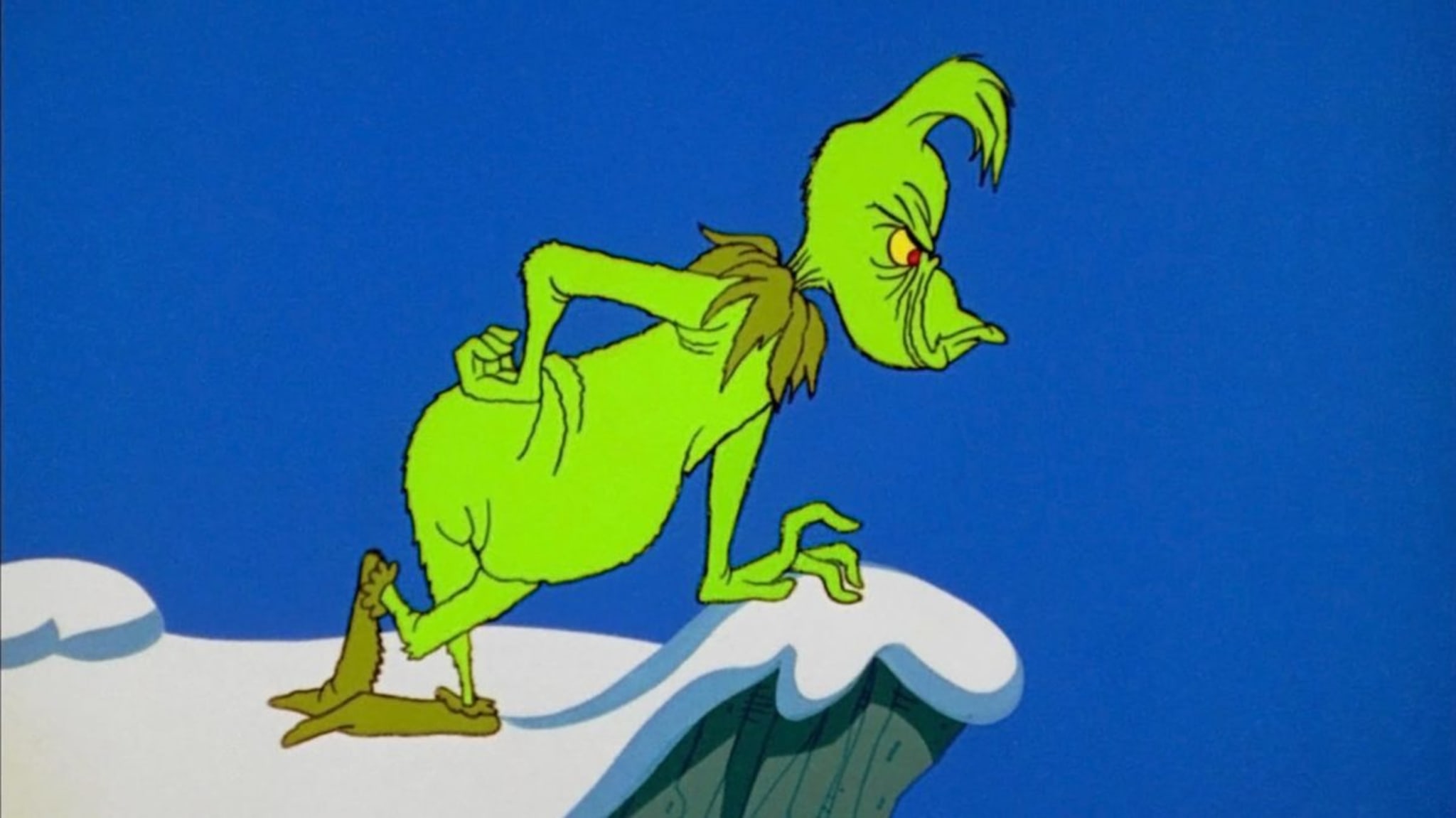 The Grinch, a green, grumpy character, stands on a snowy cliff against a blue sky, looking mischievous.