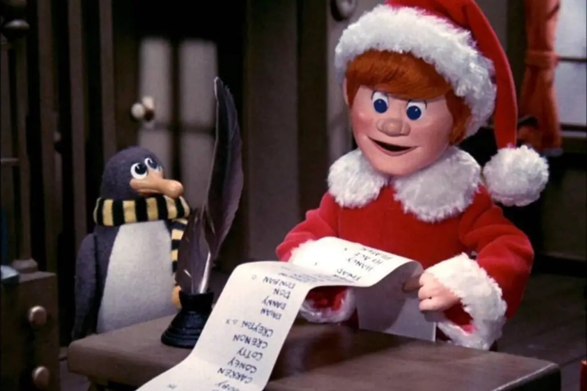 Animated scene with a character in a Santa outfit and a penguin looking at a list on a desk with a quill pen.