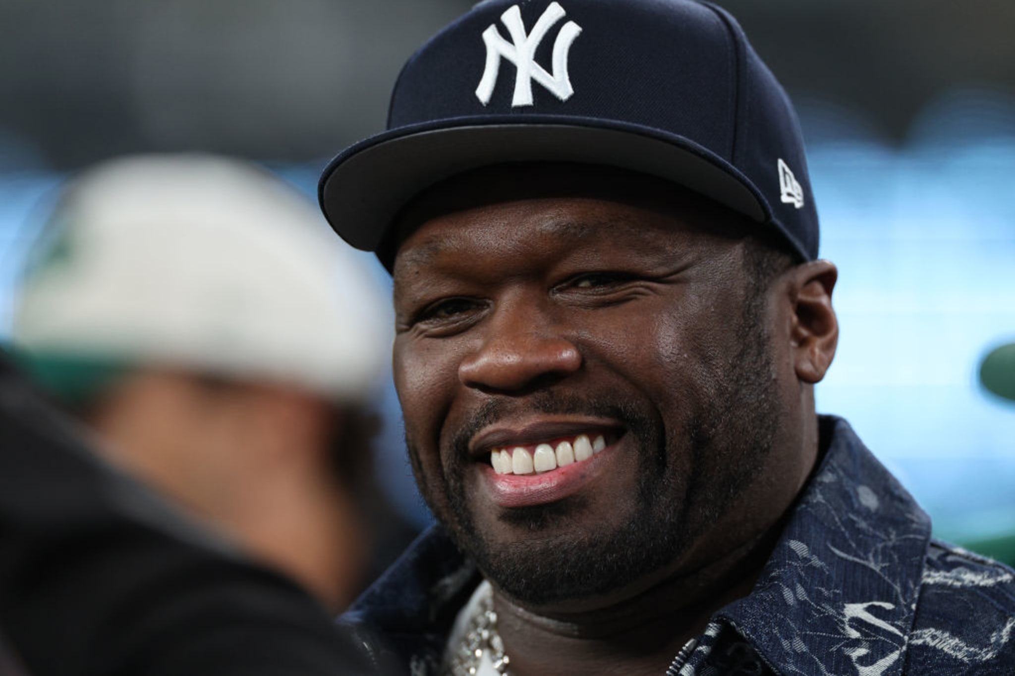 50 Cent smiling, wearing a New York Yankees cap and a patterned jacket.