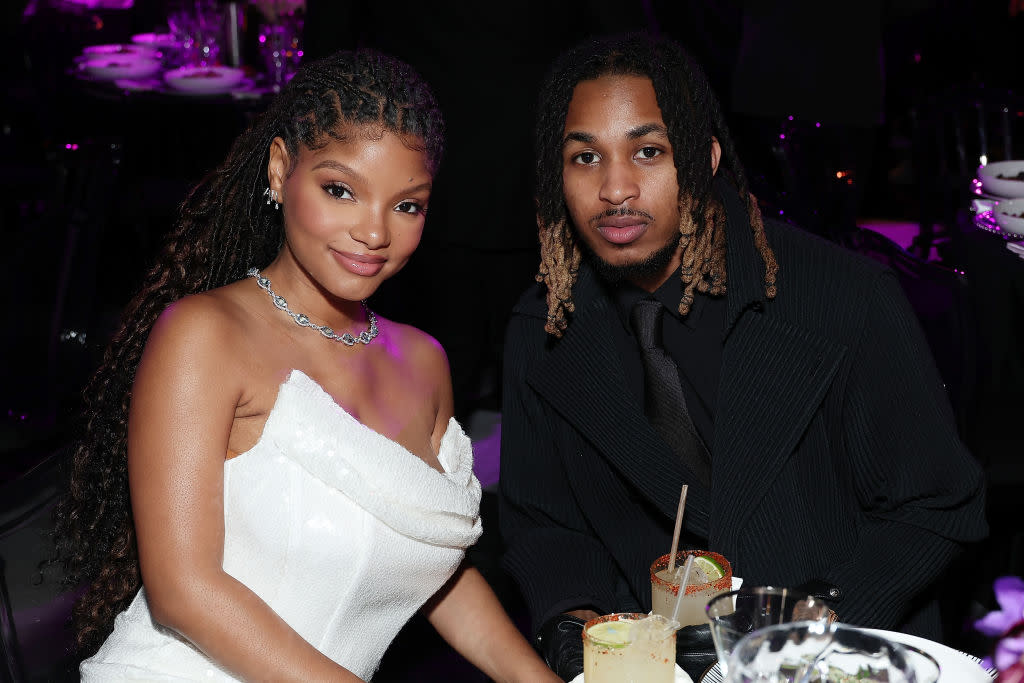 Halle Bailey in a white dress and DDG in a black suit sit together at an event, with drinks on the table.