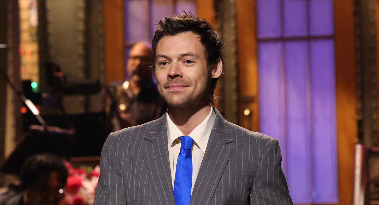 Harry Styles in a suit stands on a stage with a band in the background, likely during a live TV show.