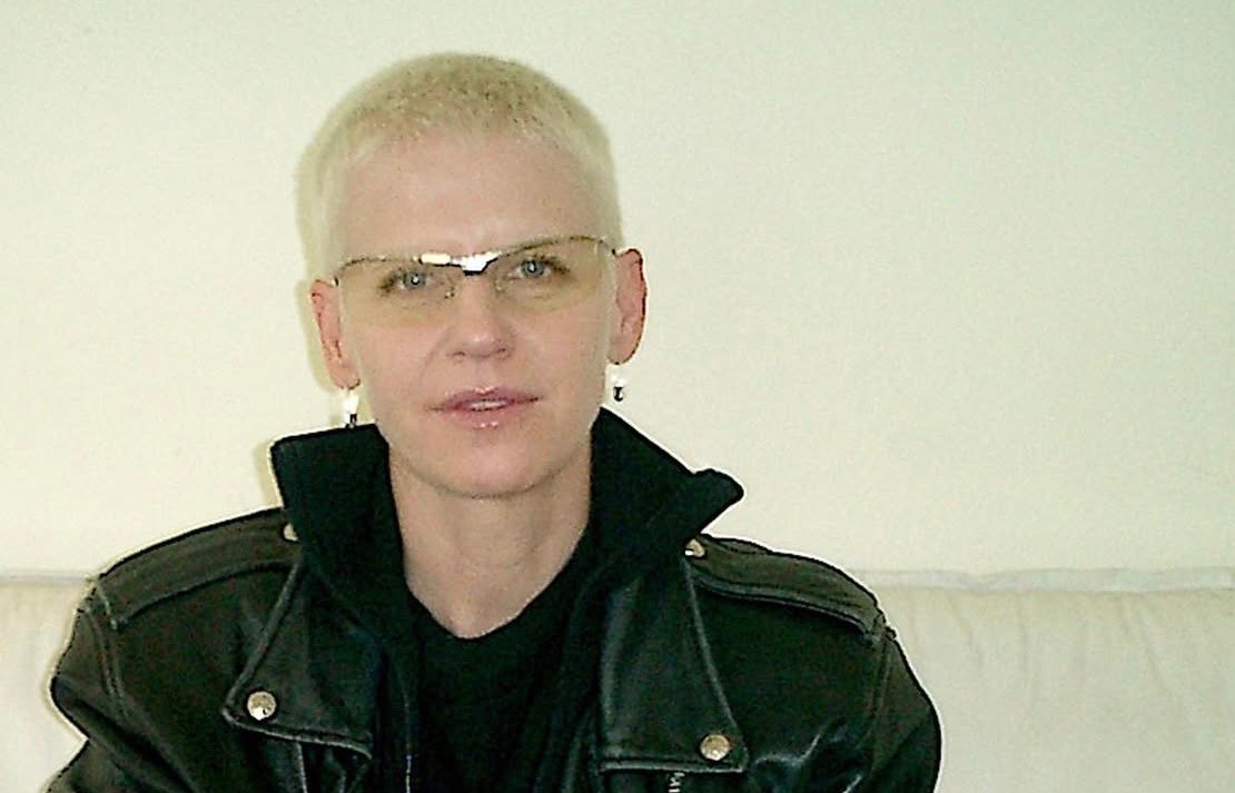 Erin Magee with short blonde hair in a leather jacket sitting on a couch, petting a black cat.
