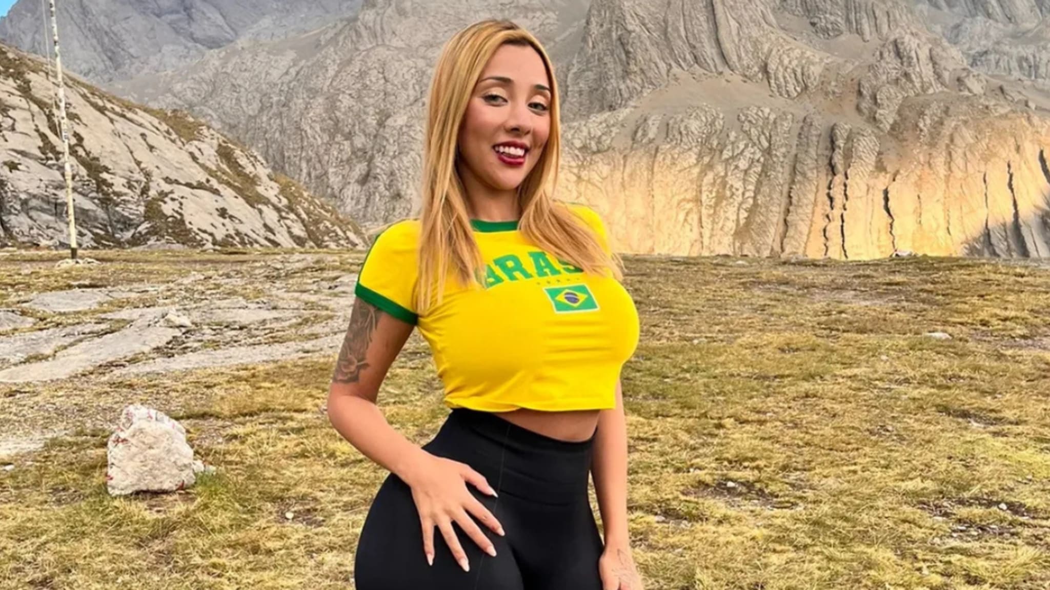 A woman in a yellow Brazil shirt and black pants poses outdoors with mountains in the background.