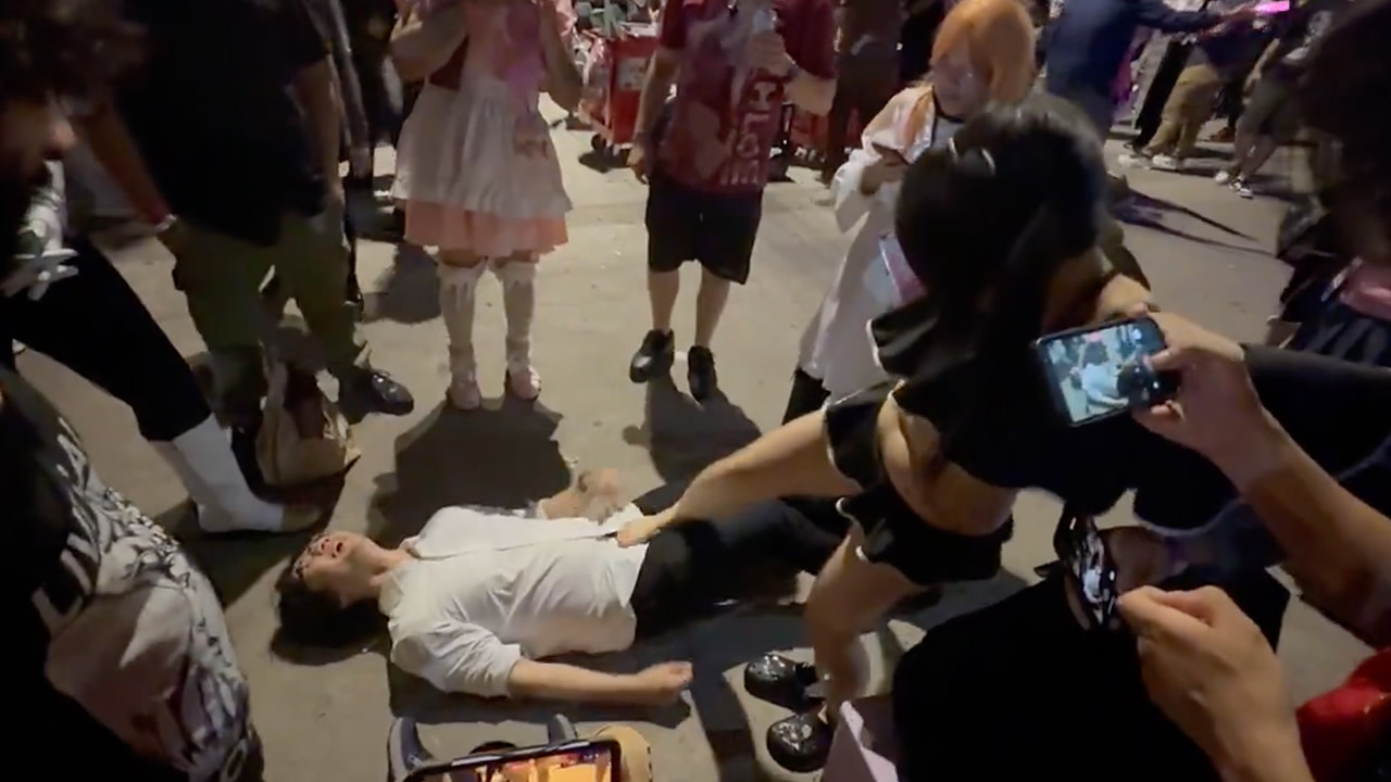 A group of people gathered around a person lying on the ground. One person is taking a photo with a smartphone.