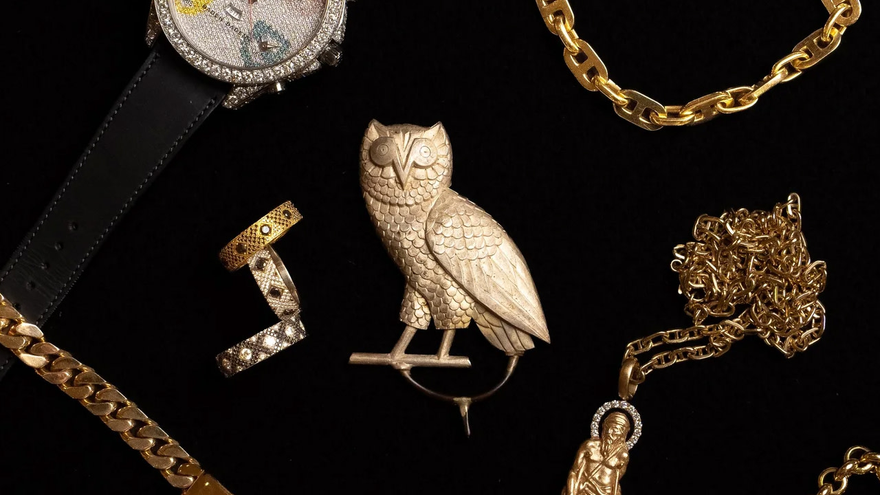 A collection of luxury items on a black background, including a diamond-encrusted watch, gold rings, a gold owl brooch, and gold chains.