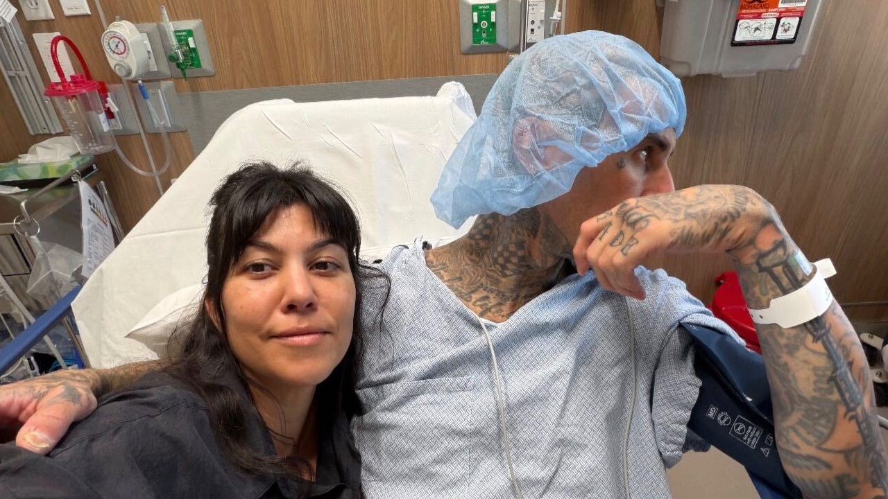 A woman and a tattooed man in a hospital gown and cap sit together in a hospital room.
