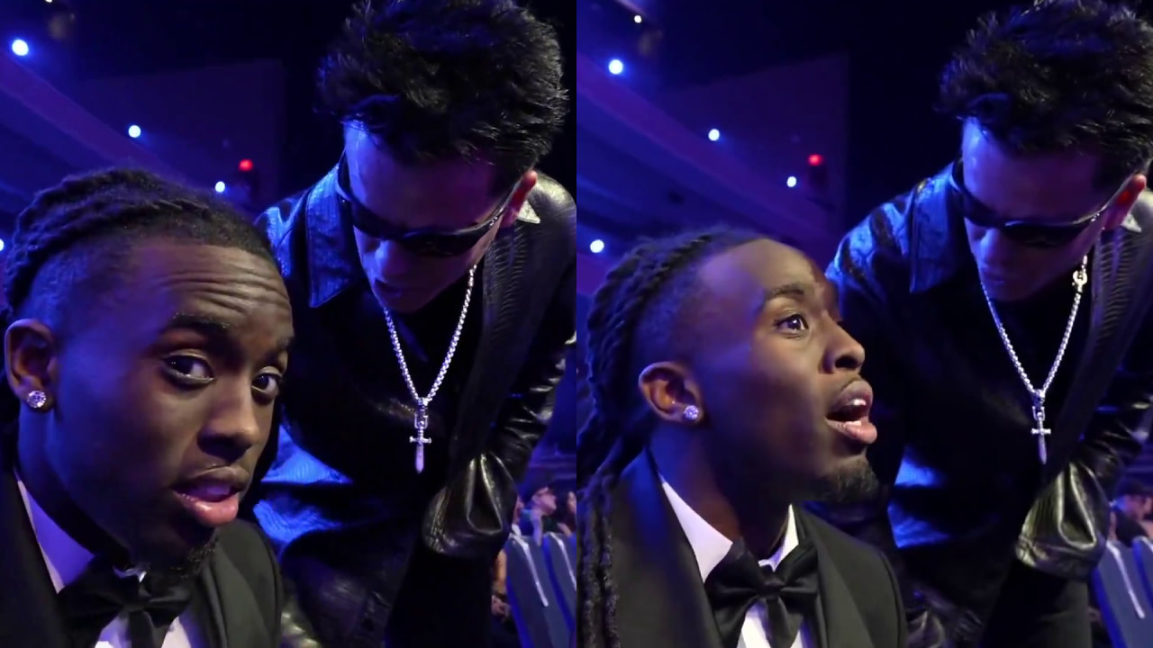 Two individuals in formal attire at an event; Kai Cenat is seated in a tuxedo, the other unknown man in sunglasses and a cross necklace, leaning in to speak.