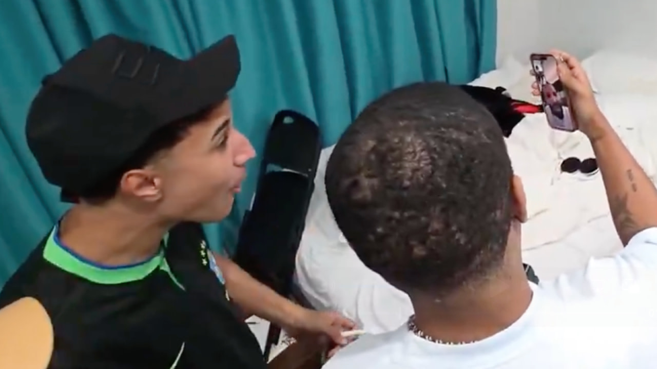 Two people taking a selfie in a room, one wearing a black cap and the other holding a phone. A bed and blue curtain are visible.