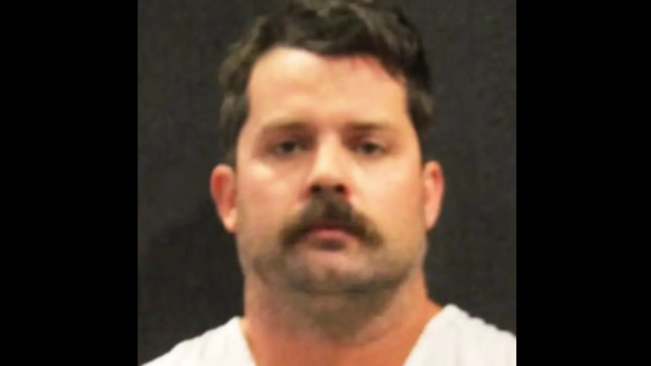 Christopher Scholtes with short dark hair and a mustache, wearing a white shirt, against a dark background.