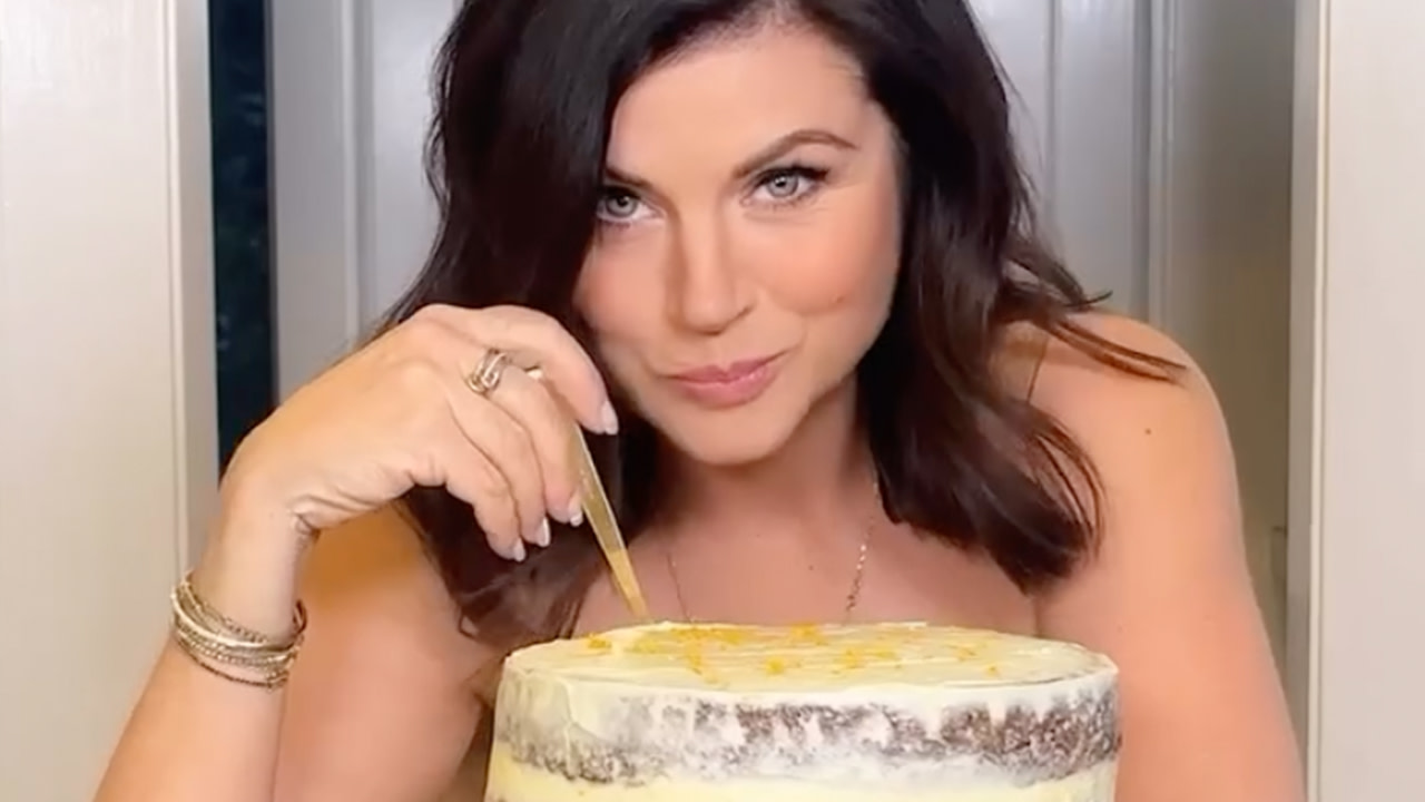 Tiffani Thiessen playfully holding a fork above a layered cake with frosting, smiling at the camera.
