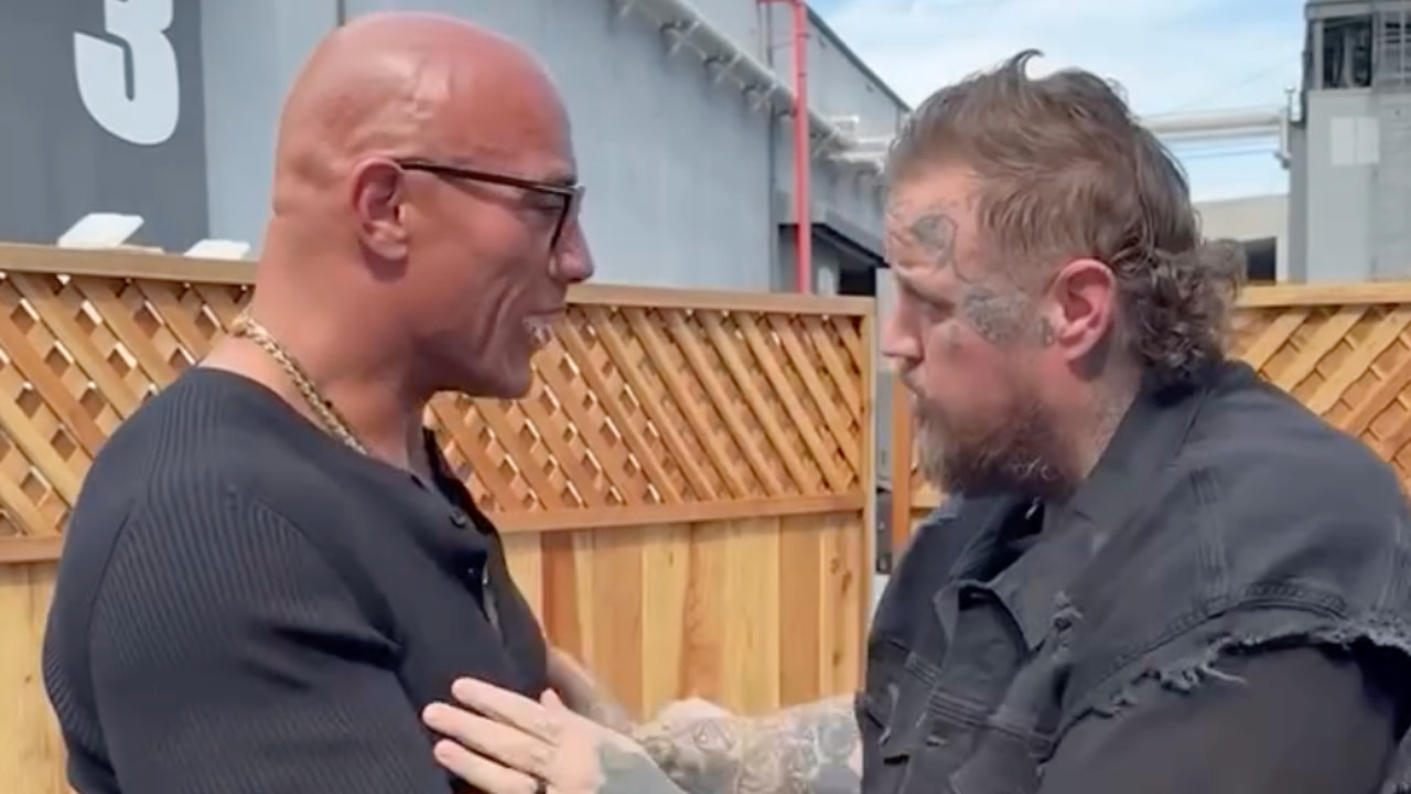 Two men facing each other, one bald with glasses, the other with tattoos and a beard. They appear to be having a conversation.