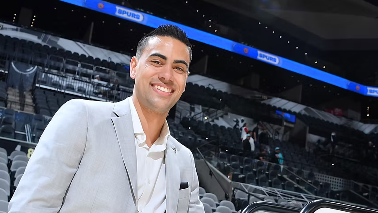 Jacob Tobey, in a light gray suit, smiles in an empty sports arena with "Spurs" on the display in the background.