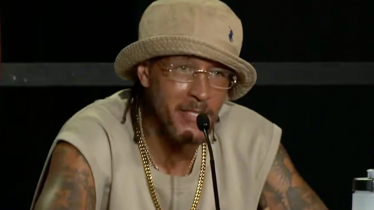 Delonte West, wearing a beige bucket hat, glasses, and a sleeveless shirt, speaks into a microphone at a panel.