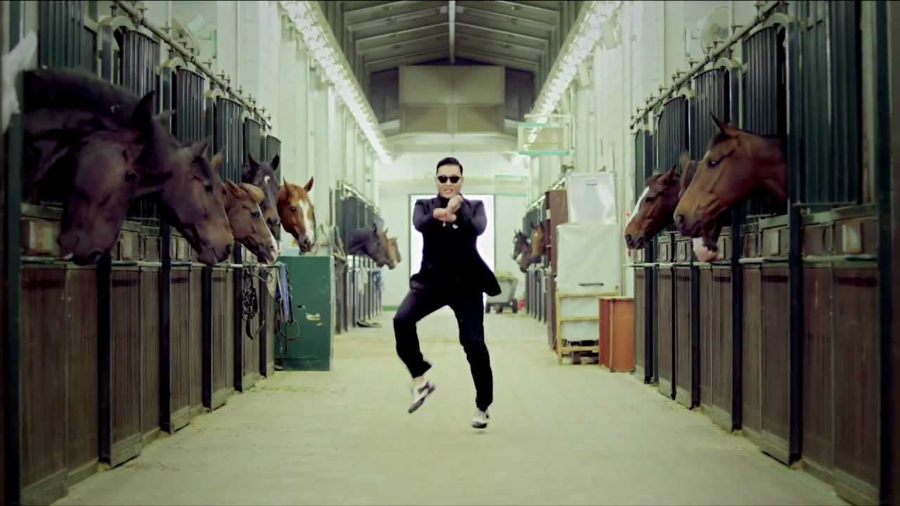 A man in a suit and sunglasses performs a dance in a stable, with horses looking out from their stalls on either side.