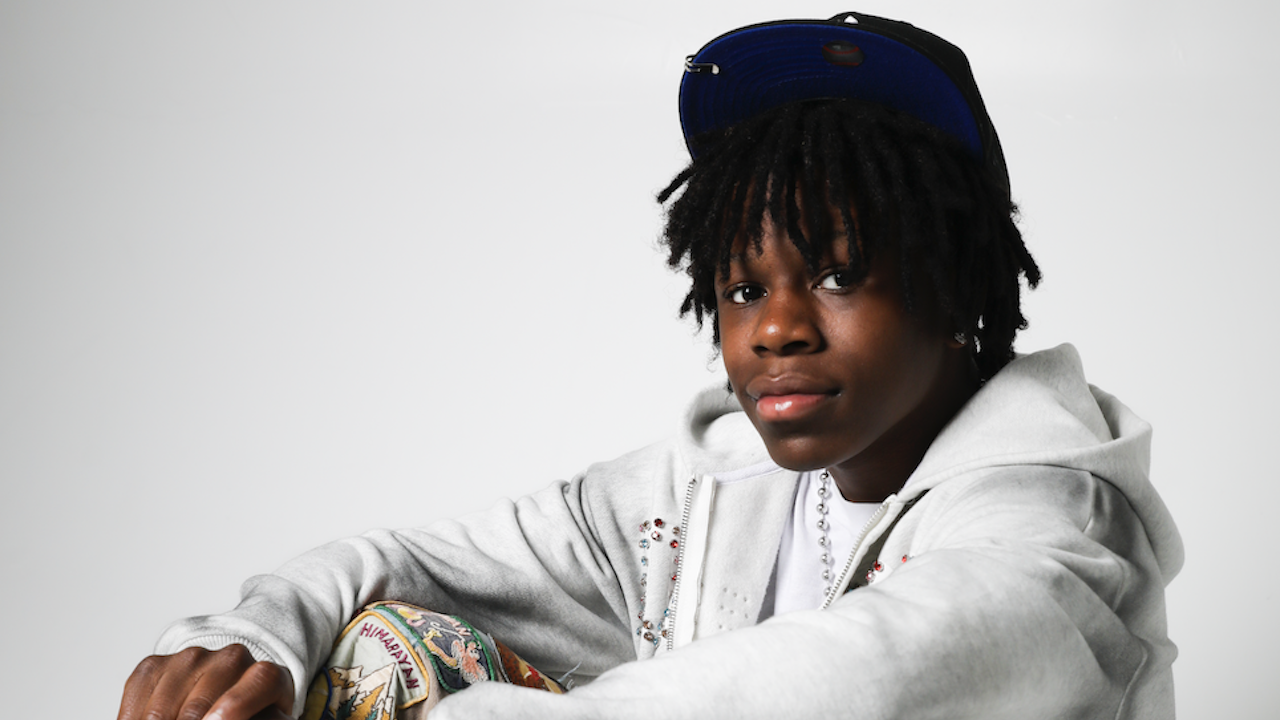 Rakai wearing a gray hoodie, colorful pants, and a cap, sitting against a white background.