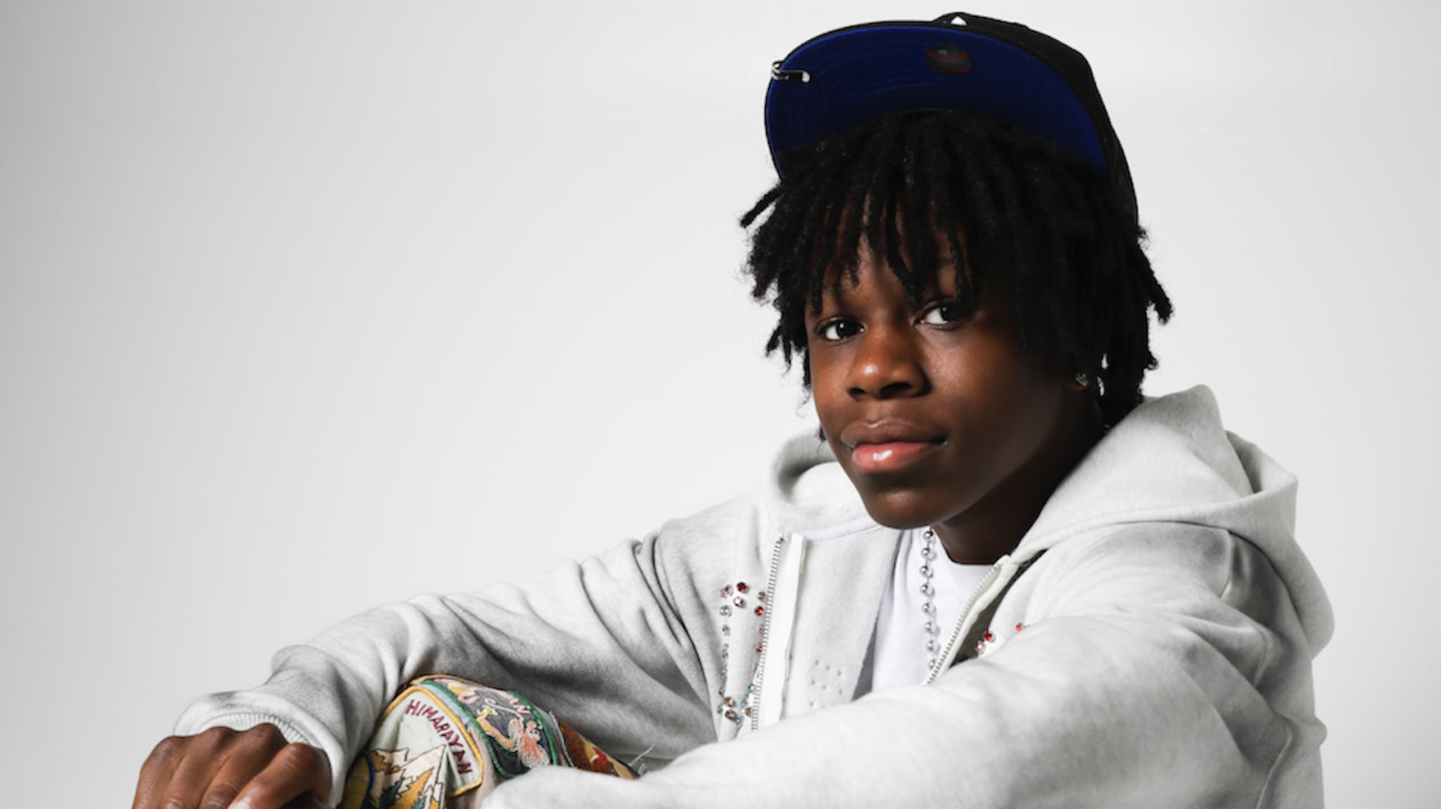Rakai wearing a gray hoodie, colorful pants, and a cap, sitting against a white background.