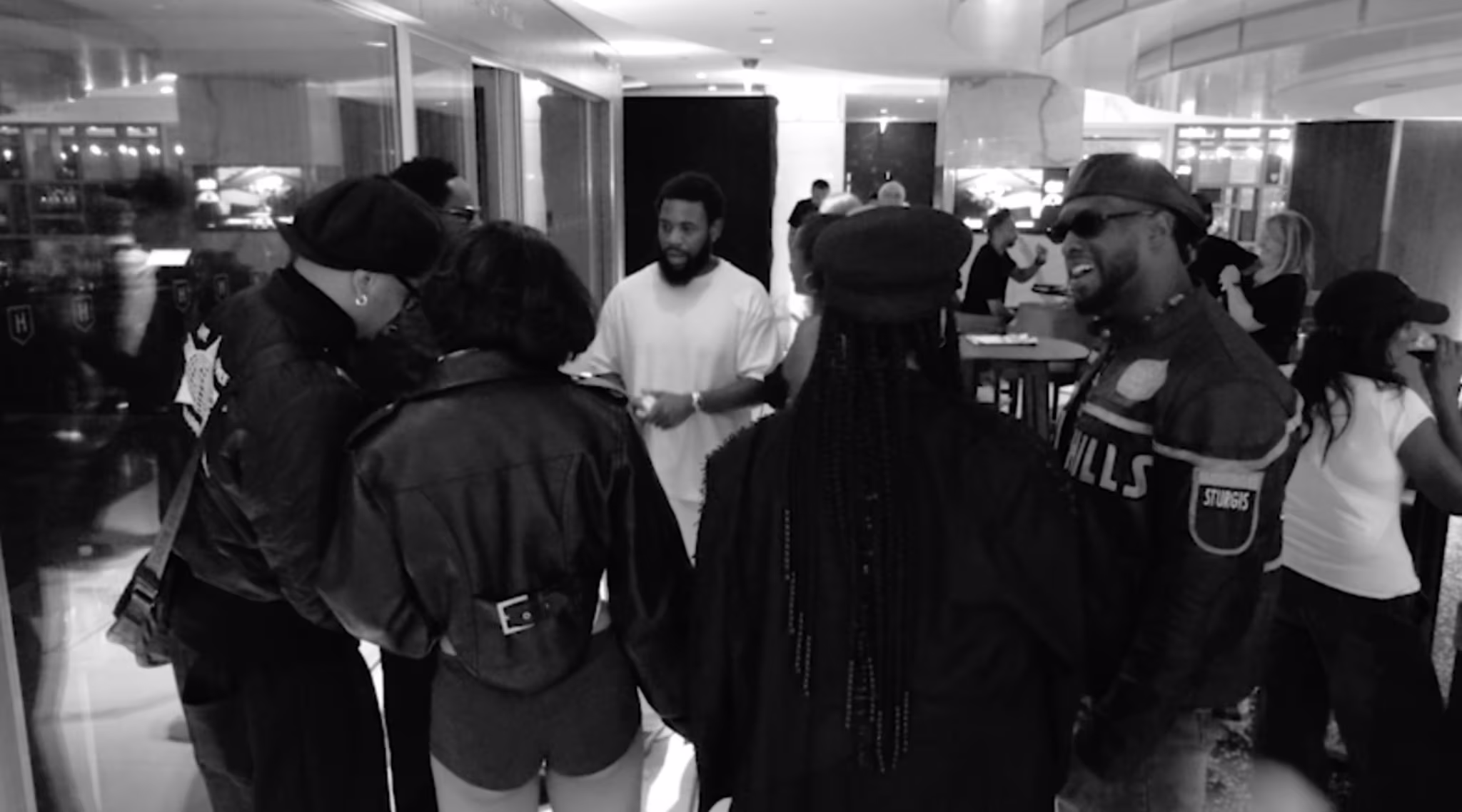 A group of people in a hotel lobby, dressed in stylish black outfits, engaged in conversation. The setting is lively and social.
