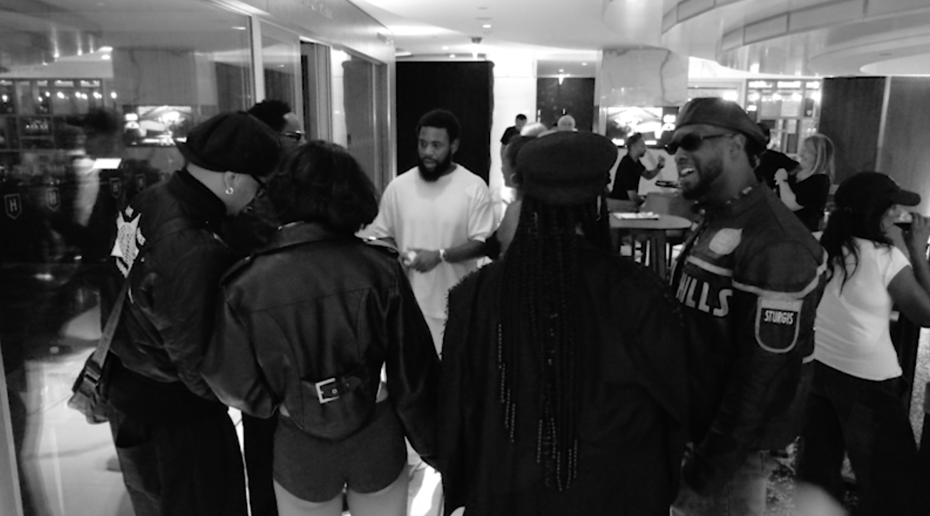 A group of people in a hotel lobby, dressed in stylish black outfits, engaged in conversation. The setting is lively and social.