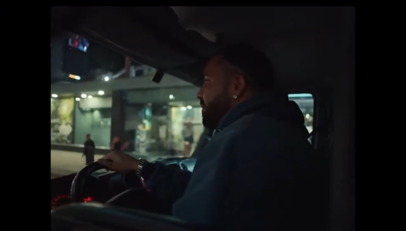 A man driving a vehicle at night, wearing a hoodie, with a city street visible outside the window.