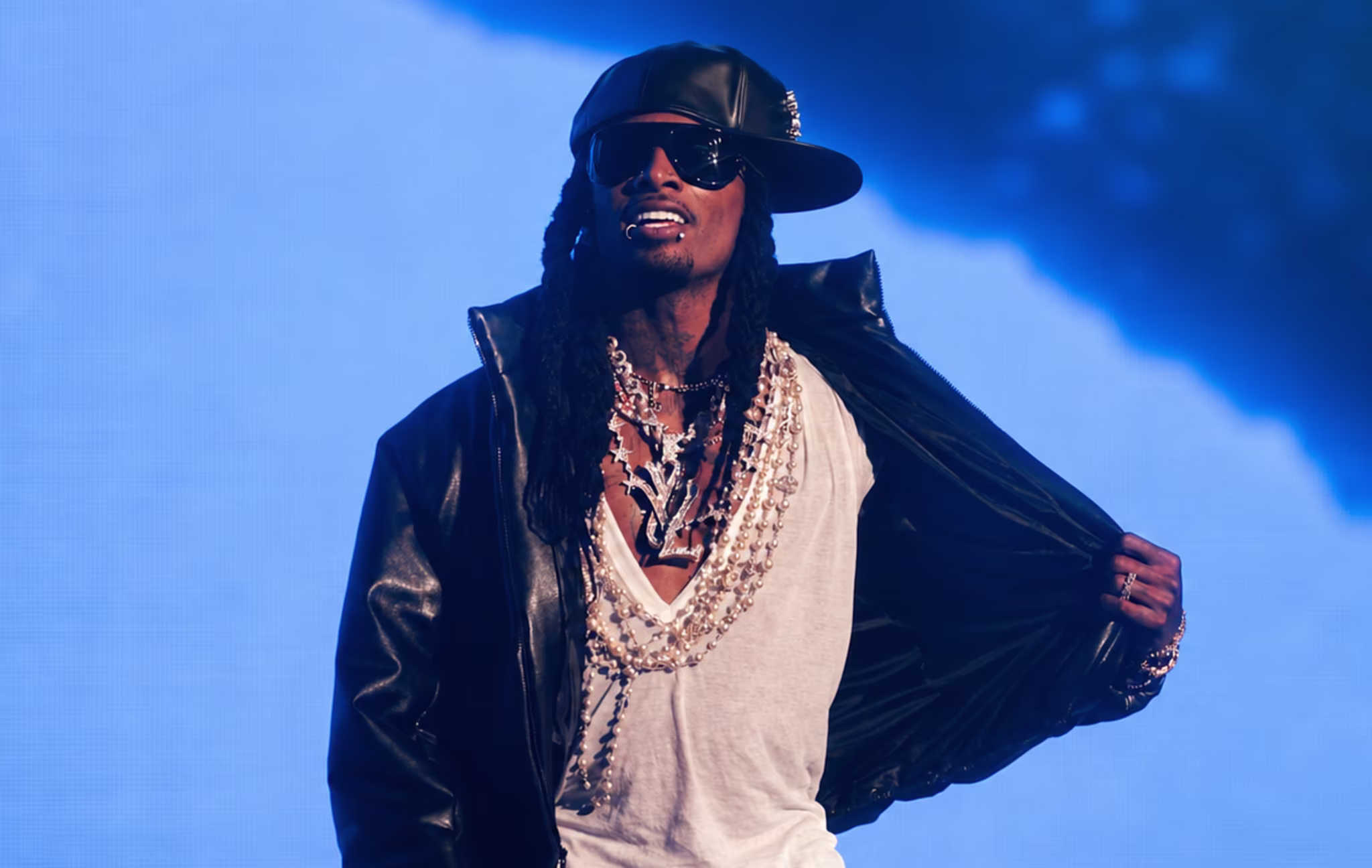 Playboi Carti wearing a black leather jacket, sunglasses, and a cap, performing on stage with a blue background.