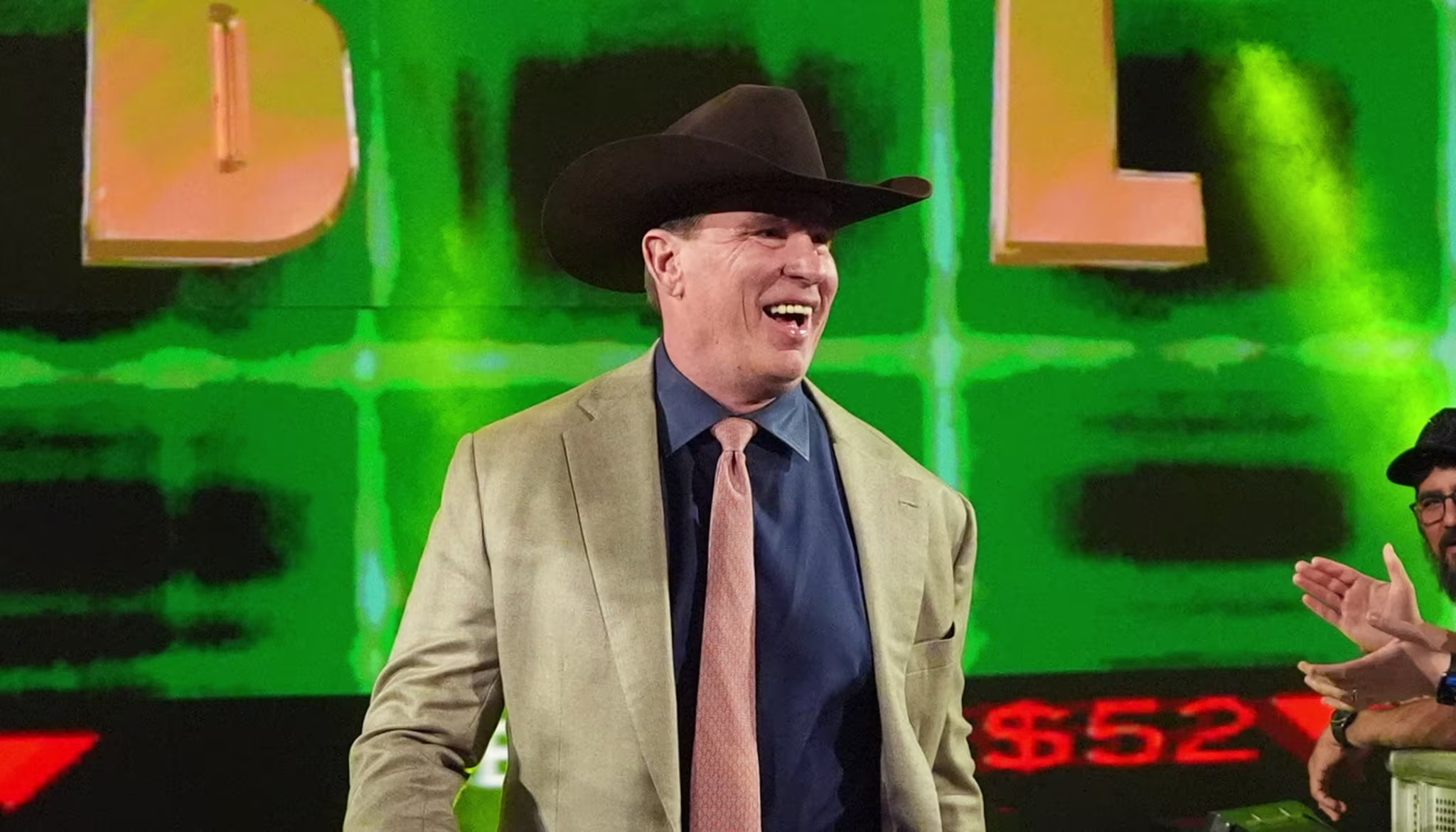 John Bradshaw Layfield in a cowboy hat and suit walks down an aisle, high-fiving fans. Large "JBL" letters are displayed on a screen behind him.