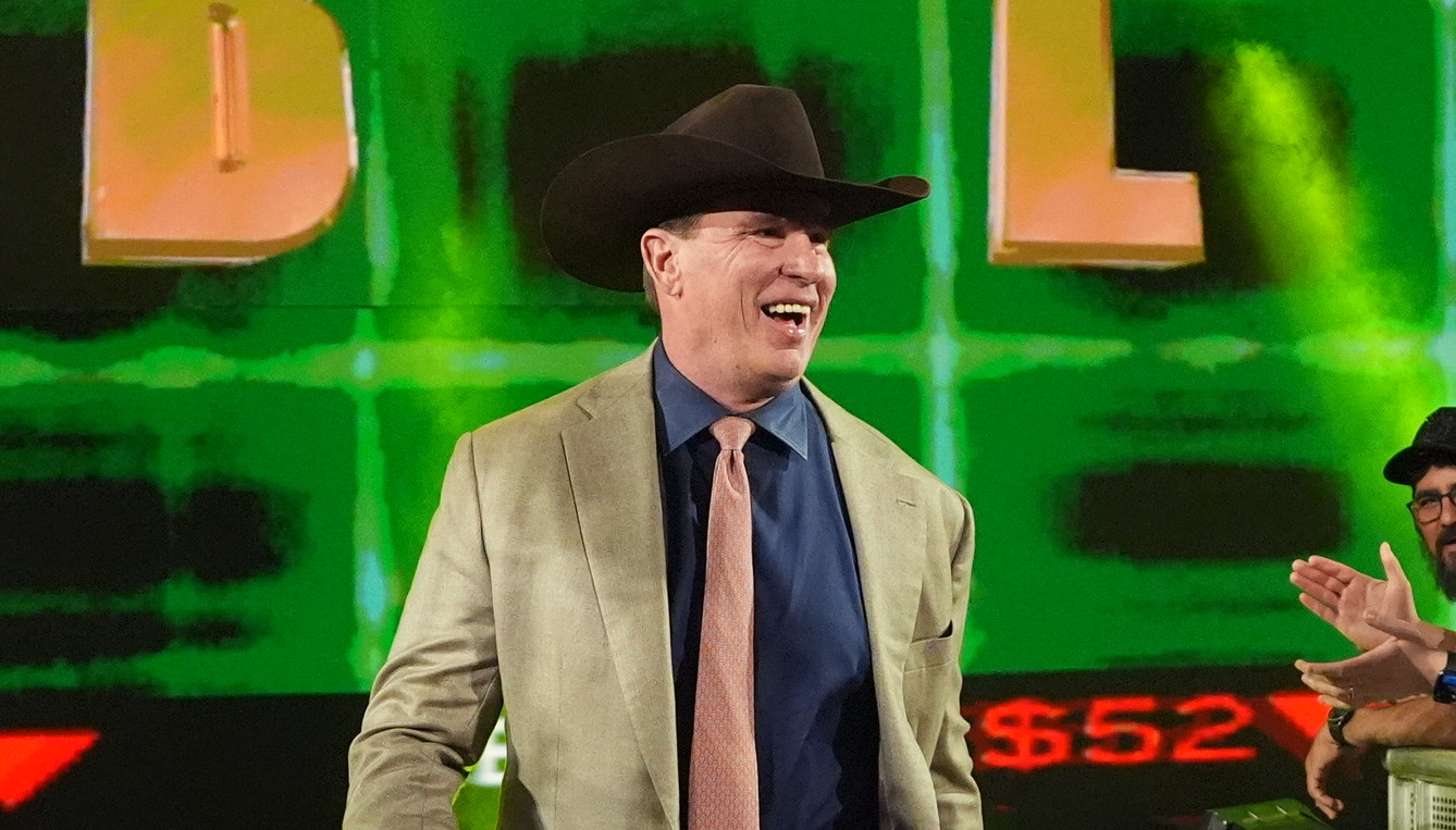 John Bradshaw Layfield in a cowboy hat and suit walks down an aisle, high-fiving fans. Large "JBL" letters are displayed on a screen behind him.