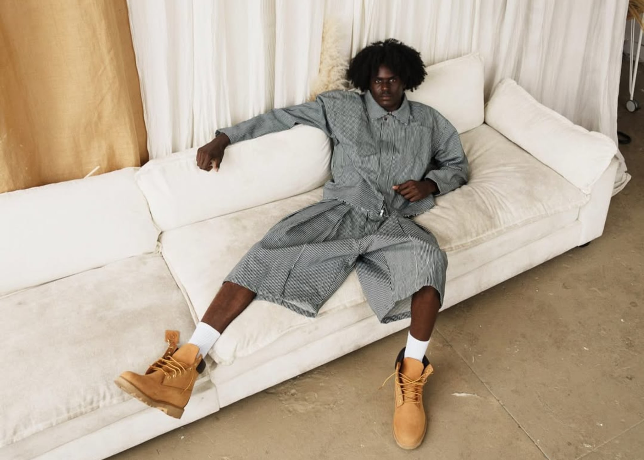 Person in a striped outfit and tan boots lounging on a white sofa, with a backdrop of beige and white curtains.