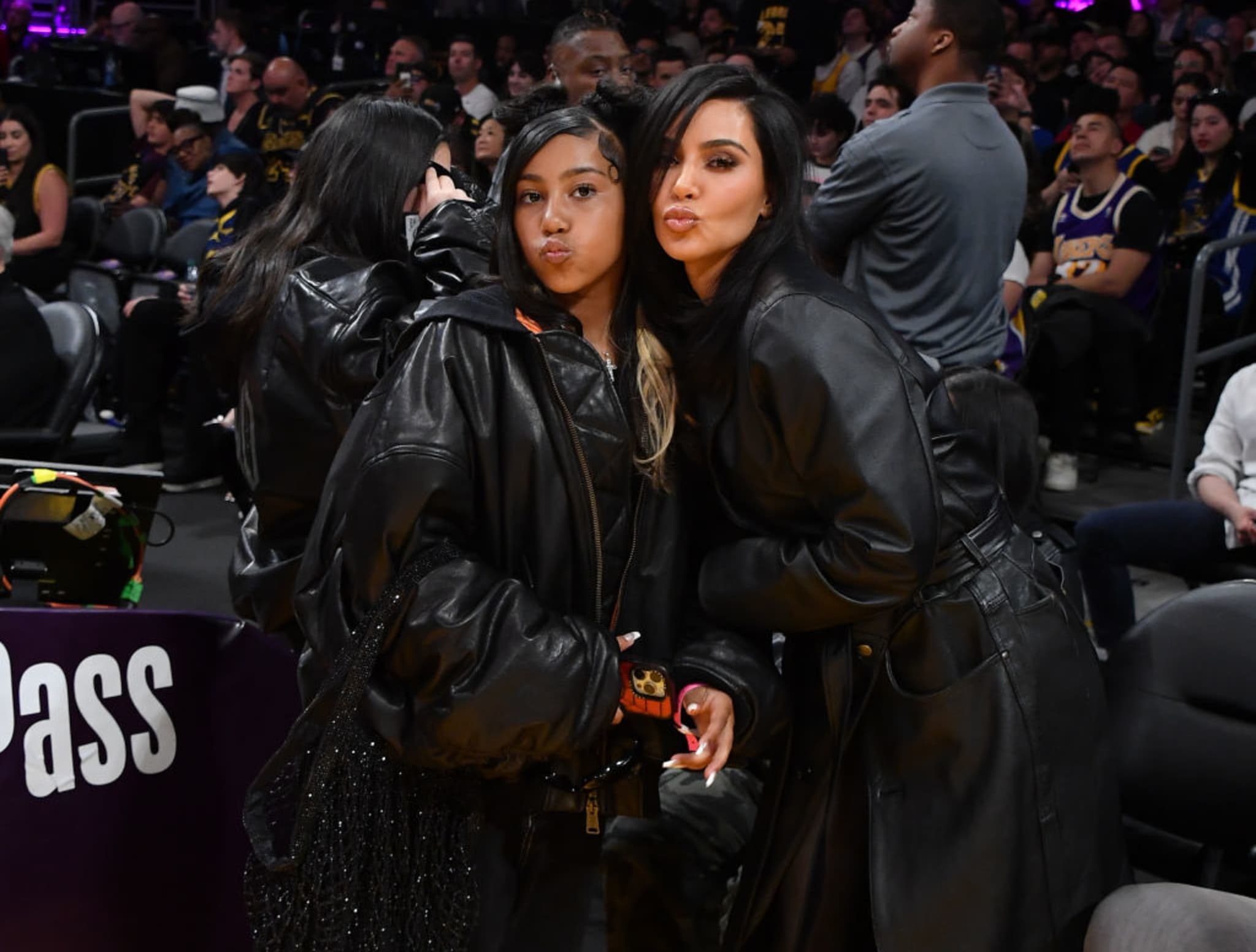Kim and North West in black outfits pose with pouting expressions at a sports event, surrounded by a crowd.