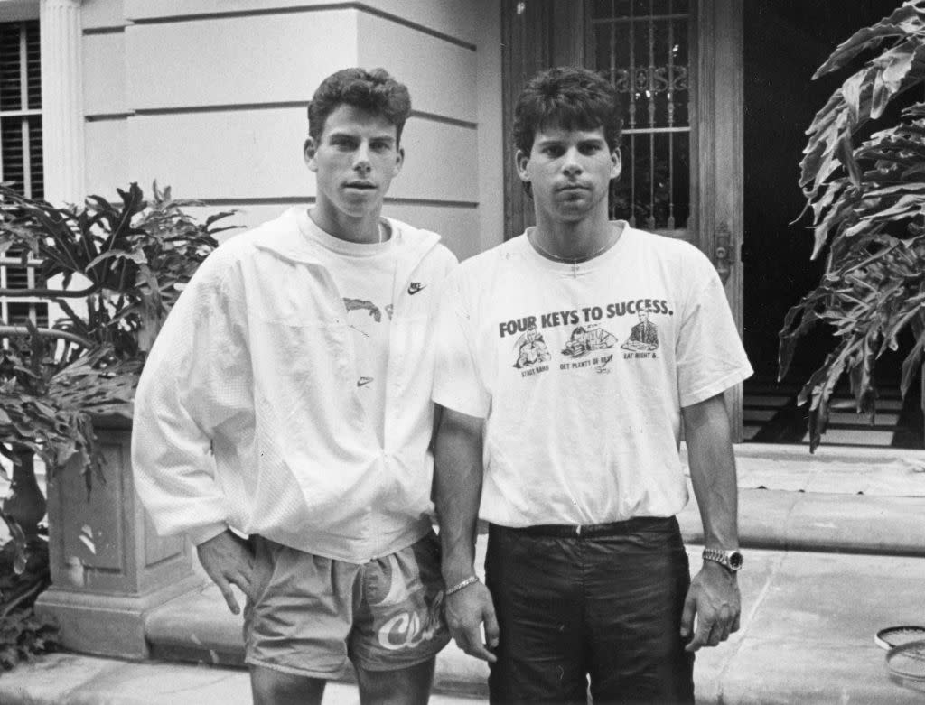 Two young men stand outside a building, one in a jacket and shorts, the other in a T-shirt and jeans, both with short dark hair.