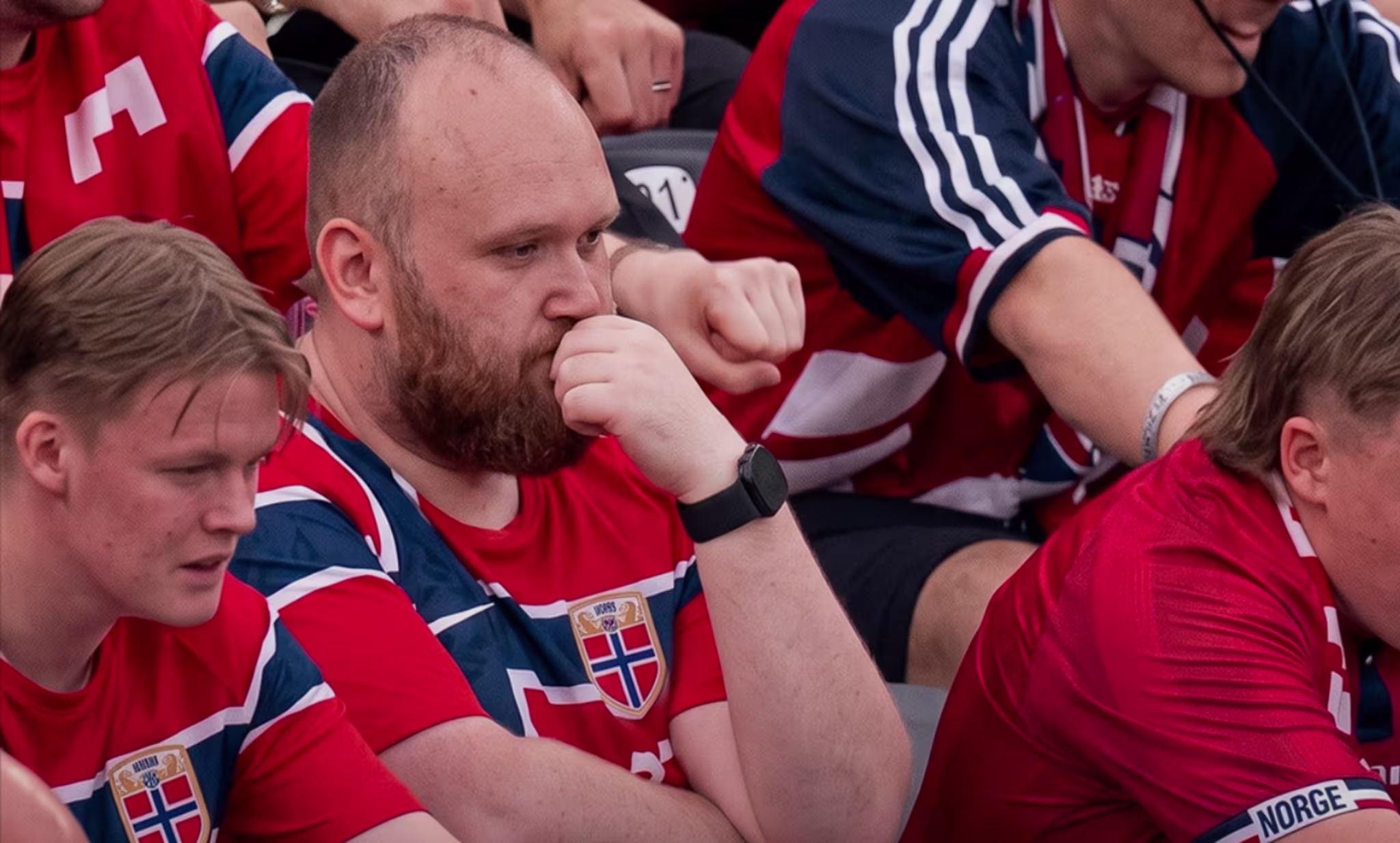 A group of people wearing red Norway jerseys, sitting in a stadium, looking focused and pensive.