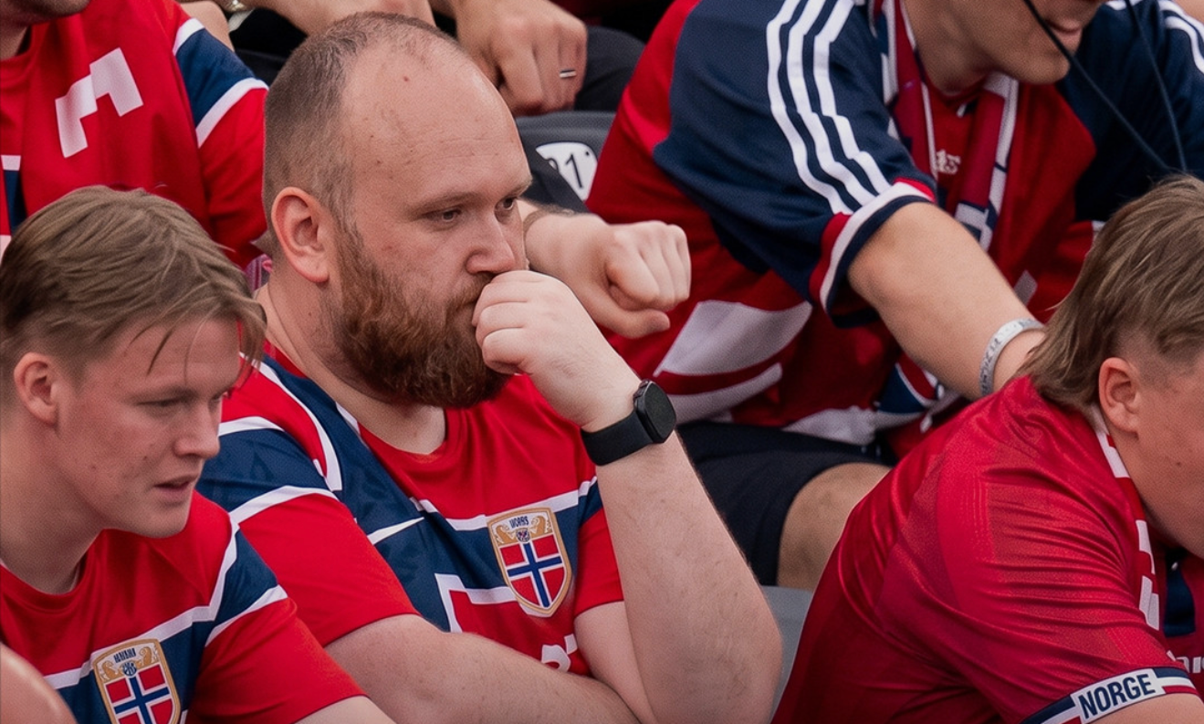 A group of people wearing red Norway jerseys, sitting in a stadium, looking focused and pensive.