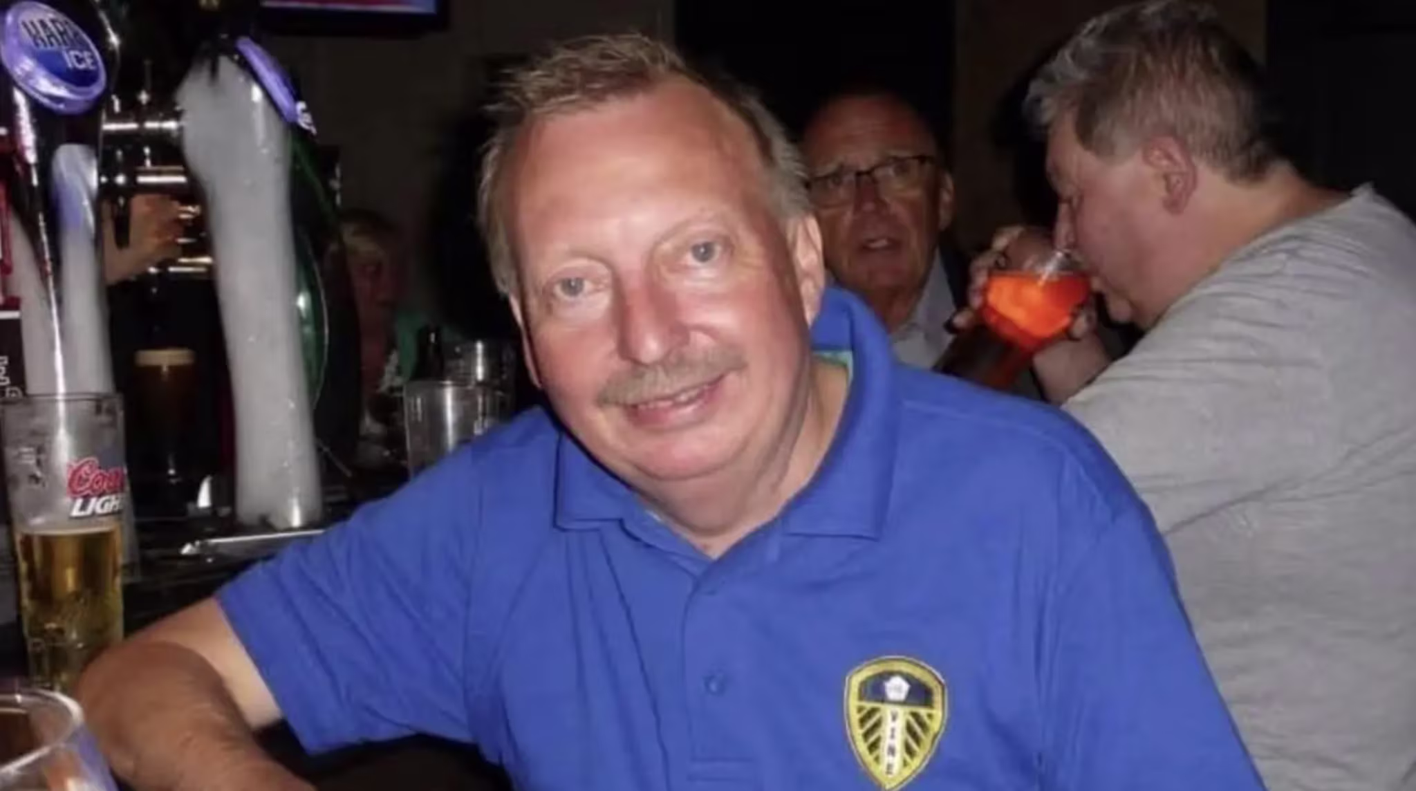 Michael Hewitt in a blue Leeds United shirt smiles at a bar, with people and drinks in the background.