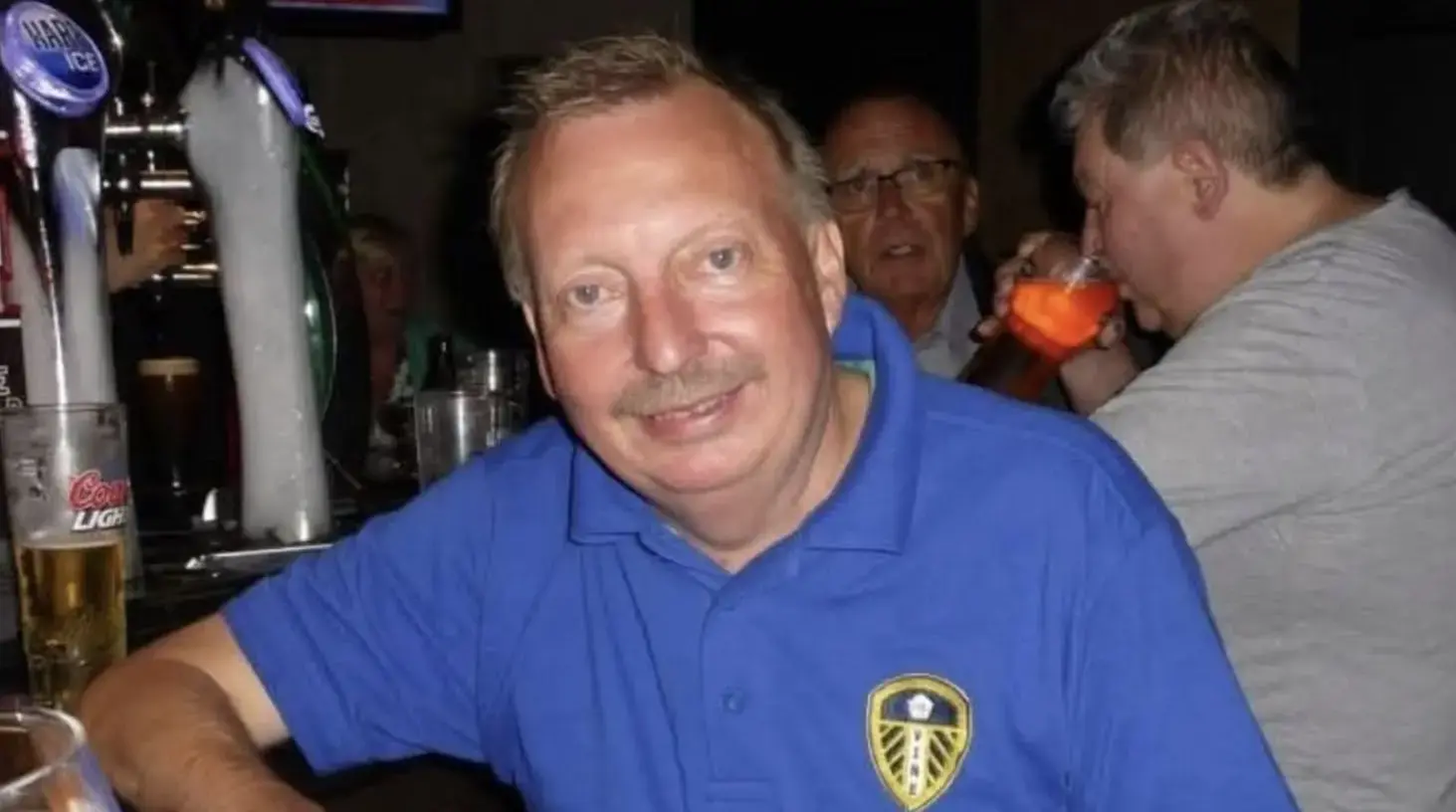 Michael Hewitt in a blue Leeds United shirt smiles at a bar, with people and drinks in the background.