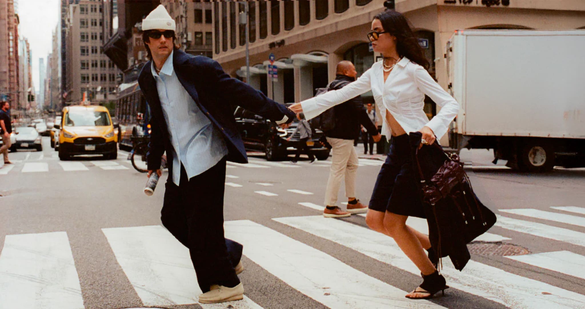 A man and woman in stylish outfits cross a city street, holding hands. The scene is urban with buildings and cars in the background.