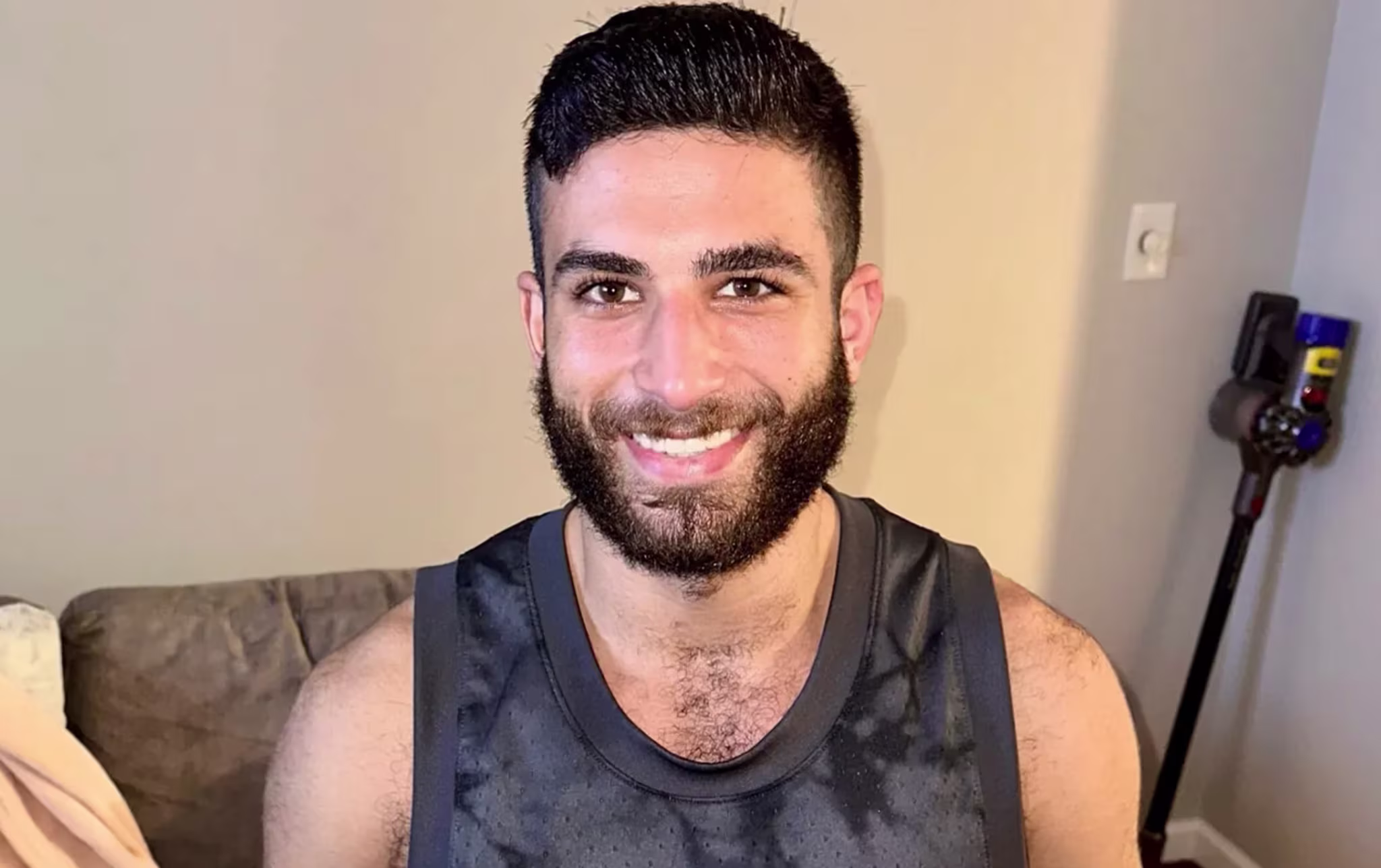 A man with a beard and short hair smiles, wearing a black sleeveless shirt. A vacuum cleaner is in the background.