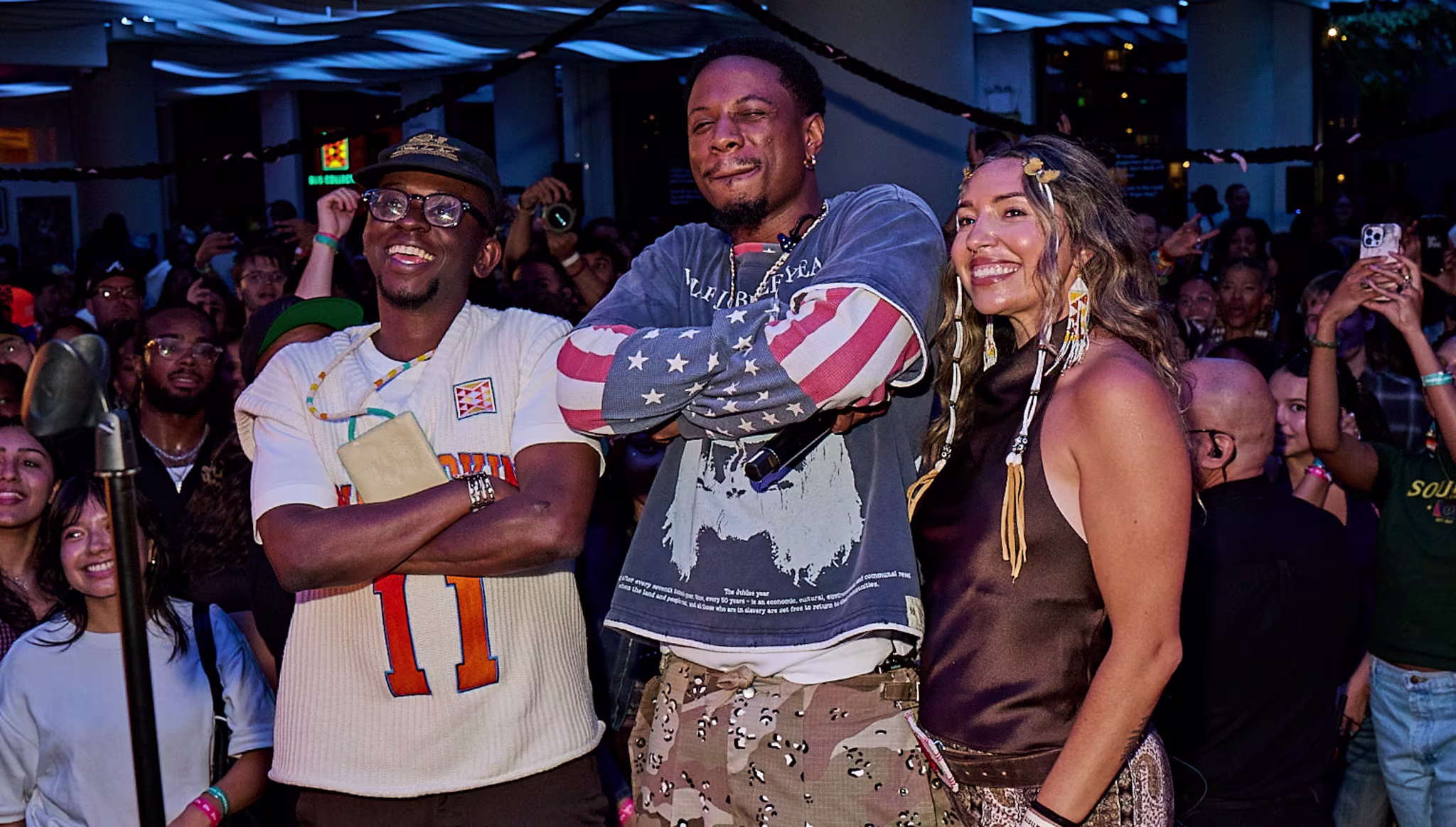 Joey Bada$$, Trevor Smith and Savannah Romero pose in front of a crowd at an indoor event with modern ceiling design and blue lighting.