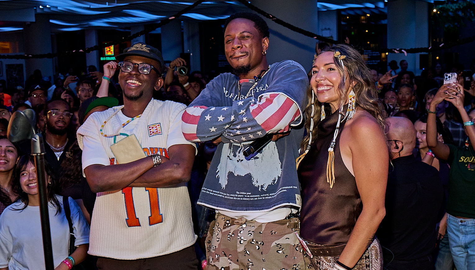 Joey Bada$$, Trevor Smith and Savannah Romero pose in front of a crowd at an indoor event with modern ceiling design and blue lighting.