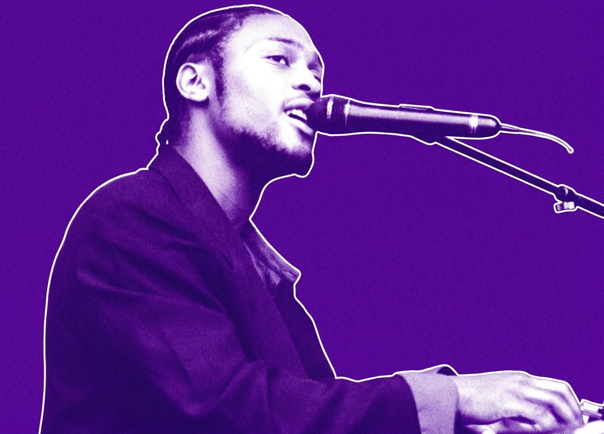 A musician with braided hair sings into a microphone while playing the keyboard, with a drummer in the background.