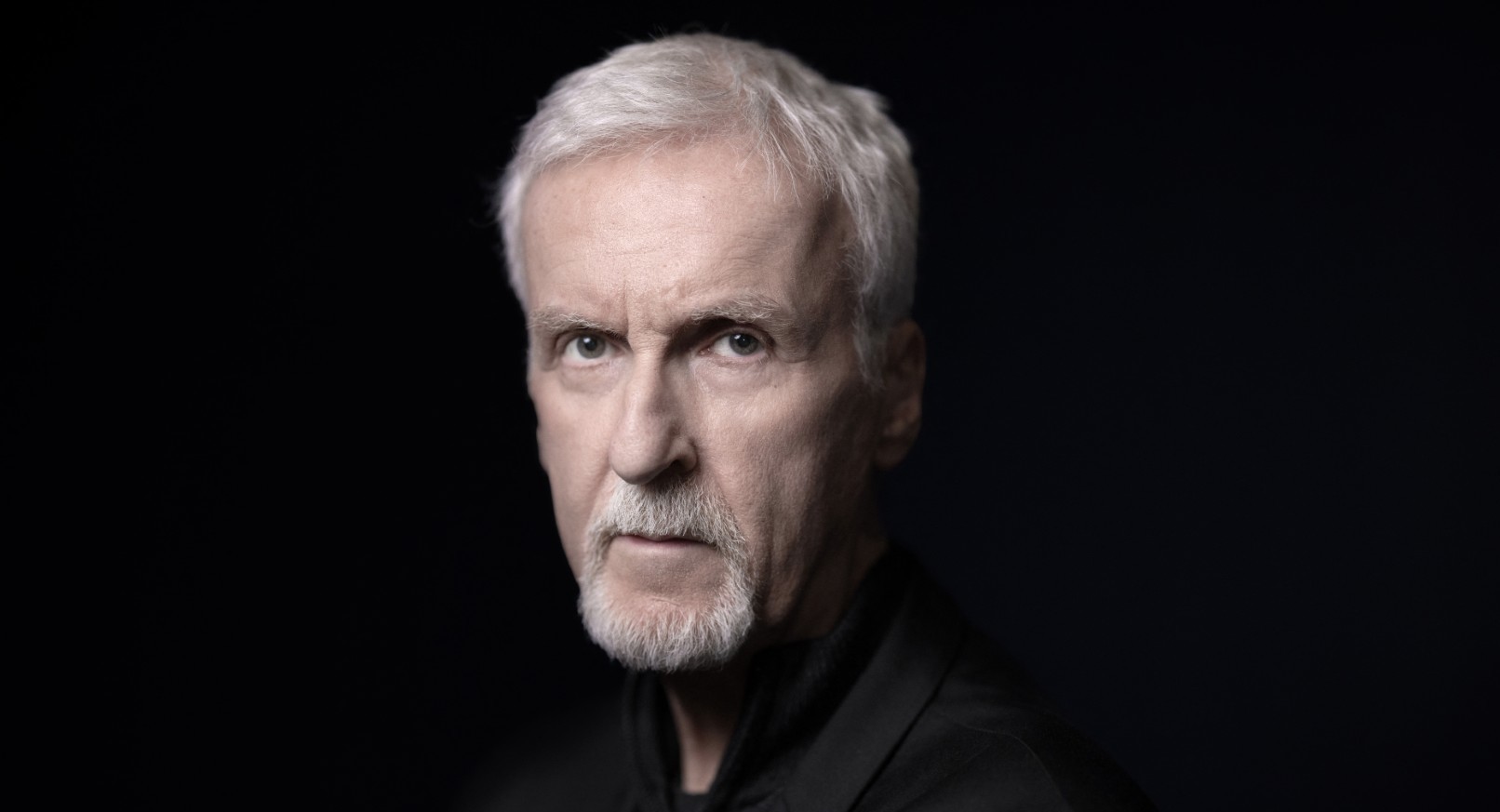 James Cameron wearing a black suit, against a dark background.