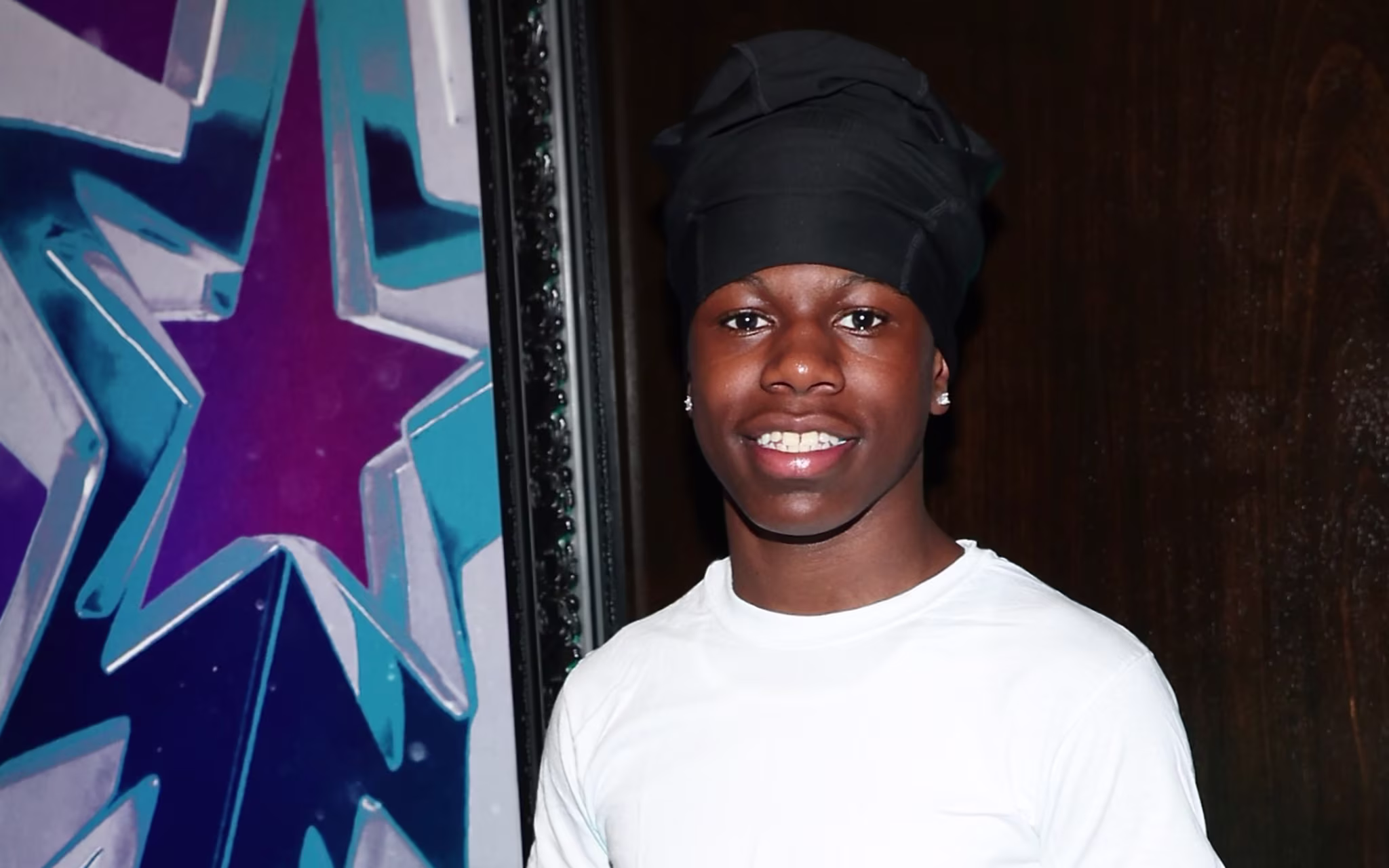 RaKai in casual attire stands beside a "BET Awards 2025" poster, smiling.