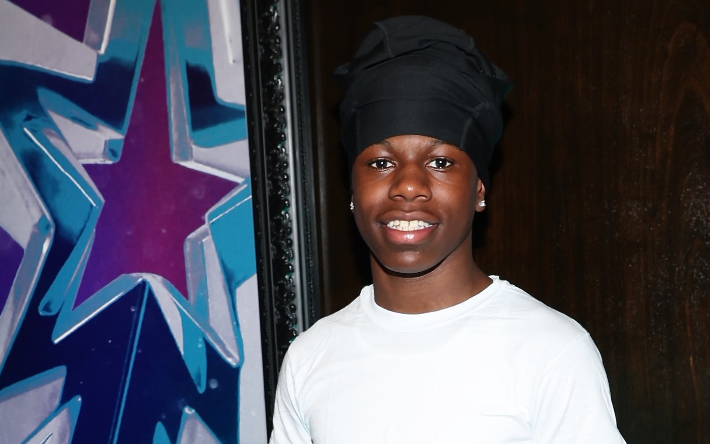 RaKai in casual attire stands beside a "BET Awards 2025" poster, smiling.
