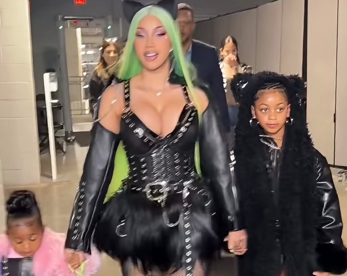 A woman with green hair in a black costume walks with two children in matching outfits, one in pink and one in black.