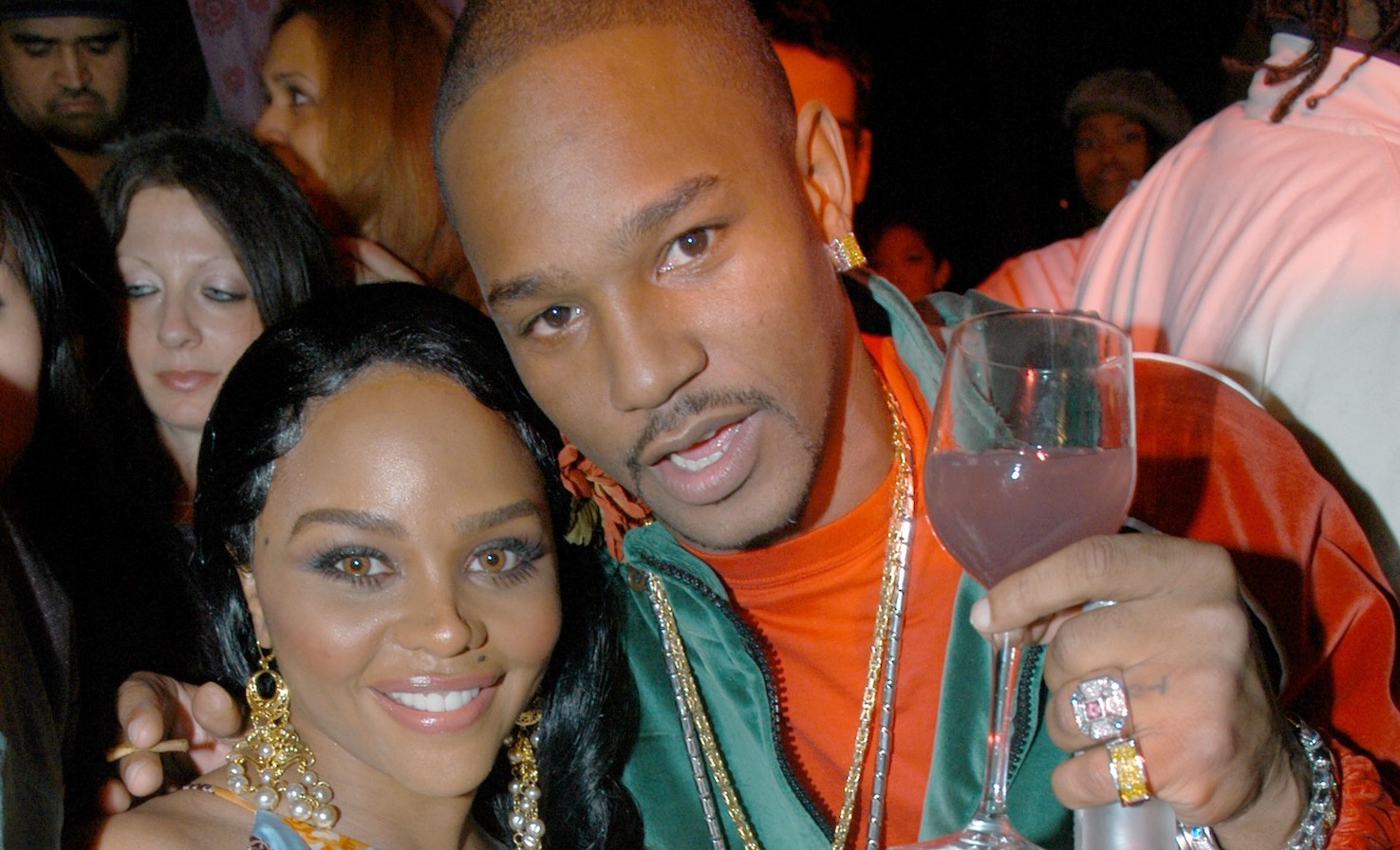Lil' Kim and Cam'ron pose together at an event, with Cam'ron holding a drink and both smiling at the camera.