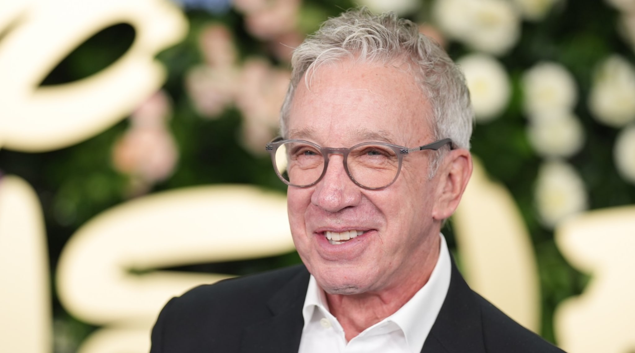 Tim Allen smiling, wearing glasses and a suit, in front of a floral backdrop.