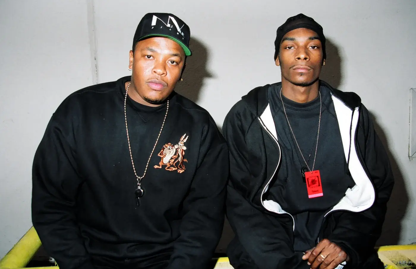 Dr. Dre and Snoop Dogg sitting together, both wearing black outfits. Dr. Dre has a black cap, and Snoop Dogg wears a beanie.
