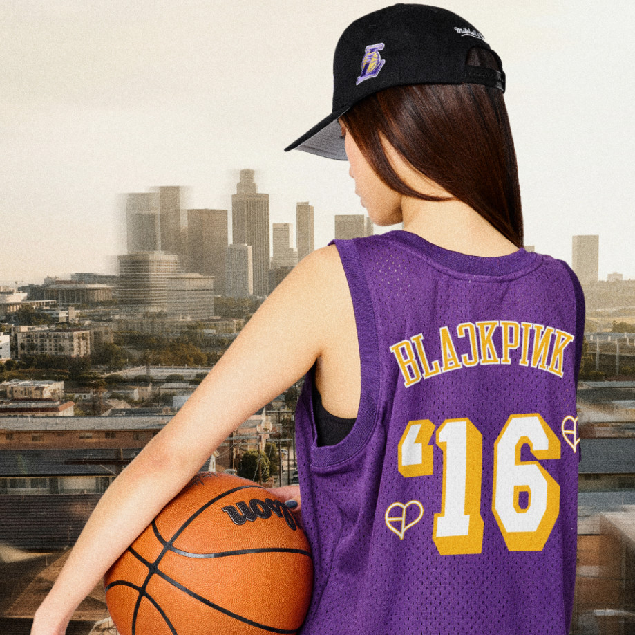 A woman in a purple jersey with "BLACKPINK '16" holds a basketball. A man in a white "Los Angeles" shirt sits, both wearing black caps.