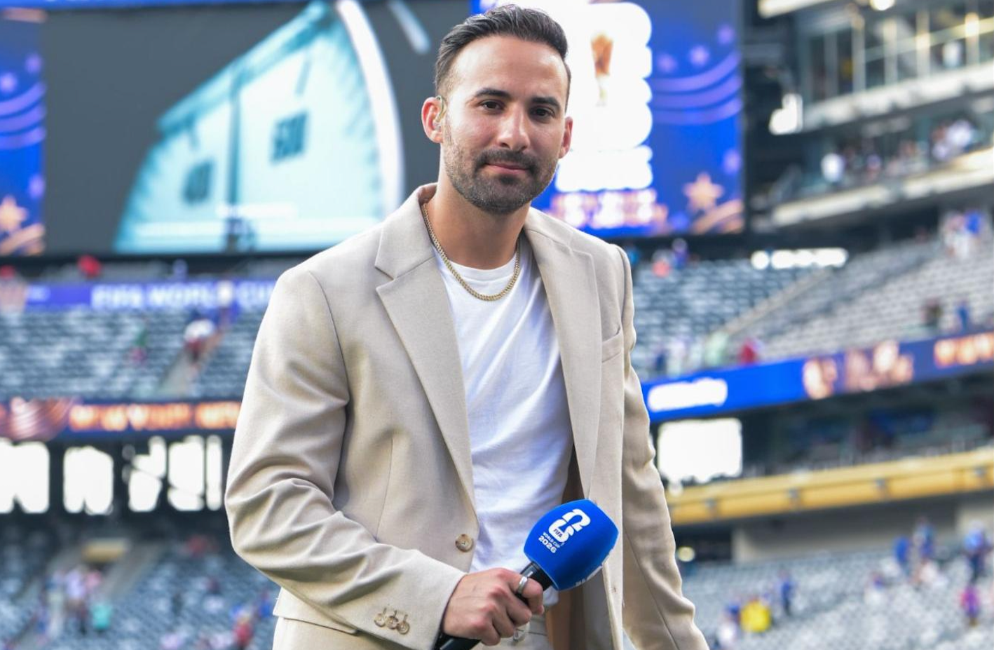 A man in a beige blazer holds a microphone in a stadium with blurred spectators and a large screen in the background.