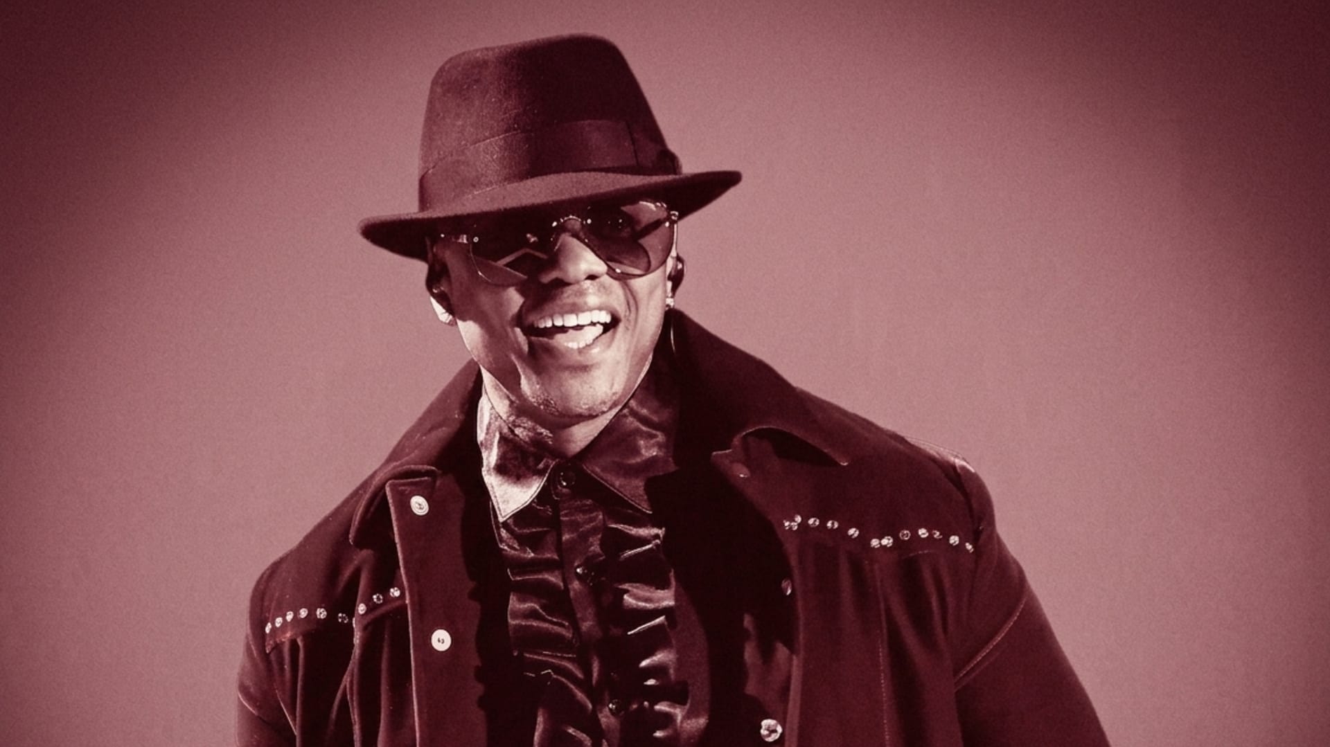 Donell Jones wearing a studded jacket, ruffled shirt, hat, and sunglasses, smiling against a sepia-toned background.