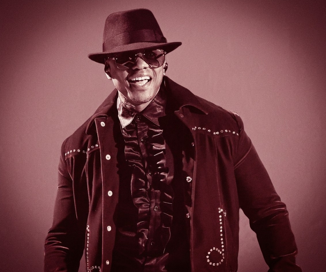 Donell Jones wearing a studded jacket, ruffled shirt, hat, and sunglasses, smiling against a sepia-toned background.